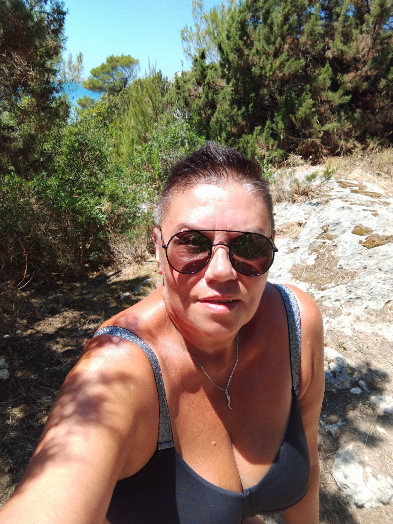 The image shows a woman taking a selfie outdoors. She has short, dark hair and is wearing aviator sunglasses and a black swimsuit with silver straps. A silver necklace with a horn-shaped pendant is visible around her neck. The woman has tanned skin, suggesting she has been in the sun. She is positioned in the foreground, with a slightly blurred background of lush green foliage, trees, and rocks. The scene appears to be in a natural, possibly coastal environment. Sunlight filters through the trees, casting shadows on the ground and the woman's arm. The sky is a clear, bright blue. The overall impression is that of a warm, sunny day, likely in a Mediterranean location, possibly Peschici, Italy, given the context. The woman's expression is neutral. No text is visible in the image.