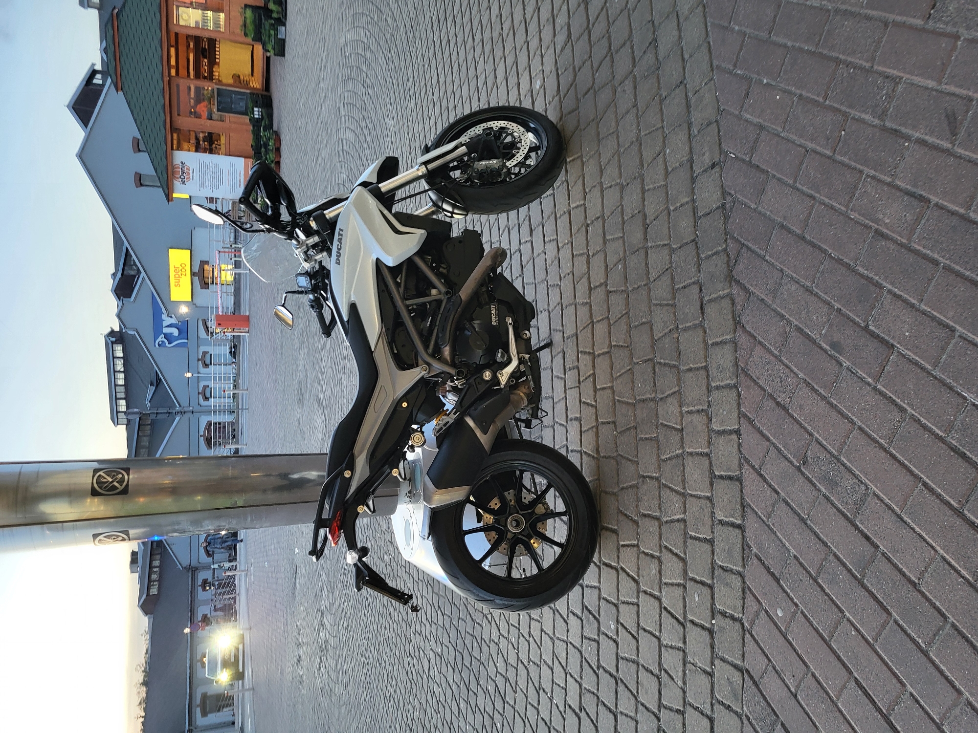 The image shows a white and black Ducati motorcycle parked on a brick and cobblestone paved area. In the background, there are buildings with visible signs in an unidentifiable language, suggesting an urban location, possibly a plaza or public space. A metal pole is near the motorcycle, and other vehicles are dimly visible in the distance. The sky suggests either dawn or dusk, given the lighting conditions.