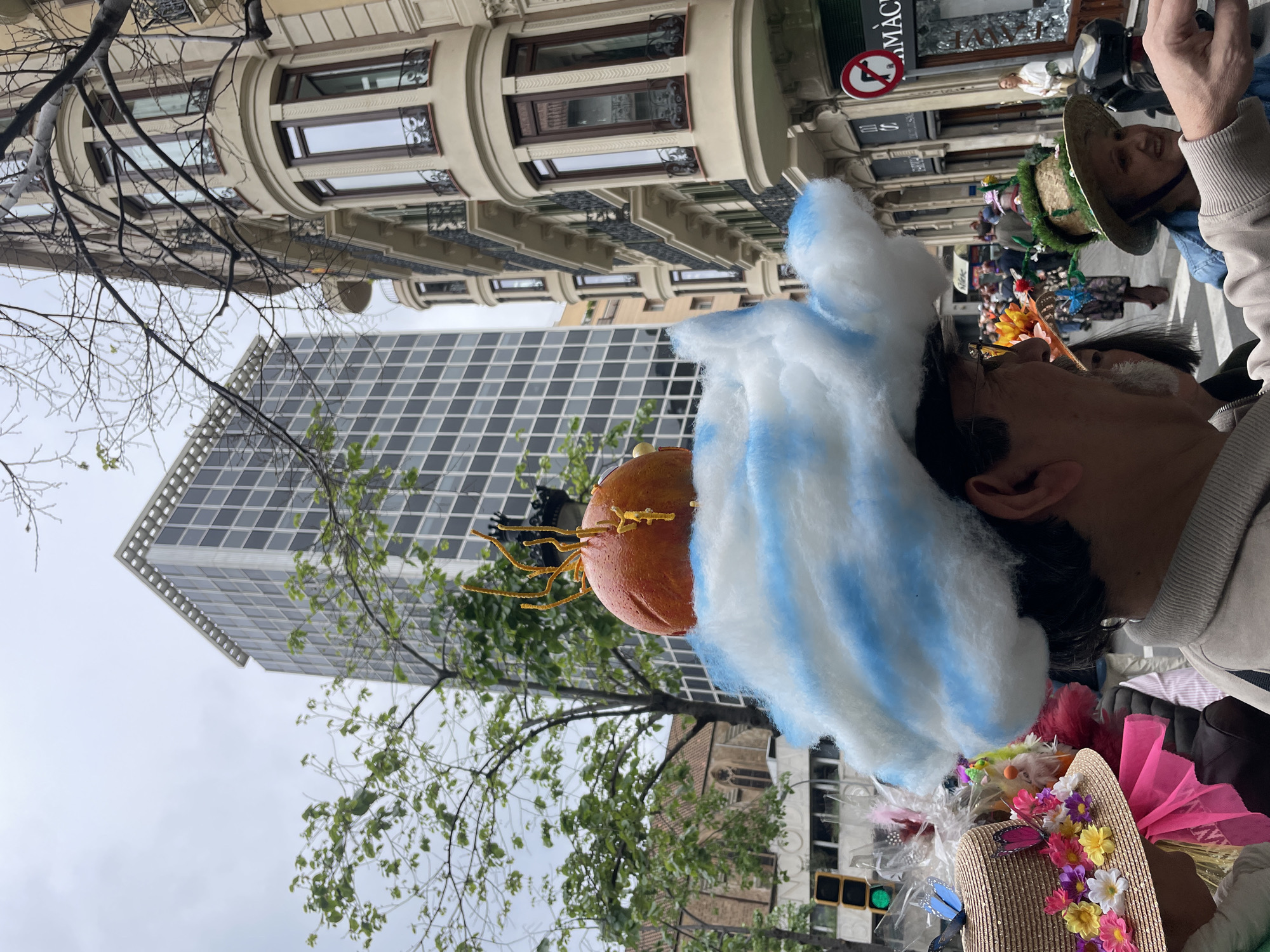 A medium eye-level shot shows a man in an Easter Parade, likely in Barcelona, Spain. He is wearing a whimsical Easter hat with a large orange sphere on top, decorated with gold-colored strands, and a large white cloud with blue stripes. In the foreground, there is a brown woven hat decorated with colorful flowers. The background contains other parade participants wearing hats. Behind the people, a modern high-rise building with a checkered pattern of windows is visible, along with a traditional building with ornate windows and balconies. A leafy green tree occupies the lower left corner. The sky is overcast. A traffic light is green. A store sign visible on the upper right corner reads "FARMAC."