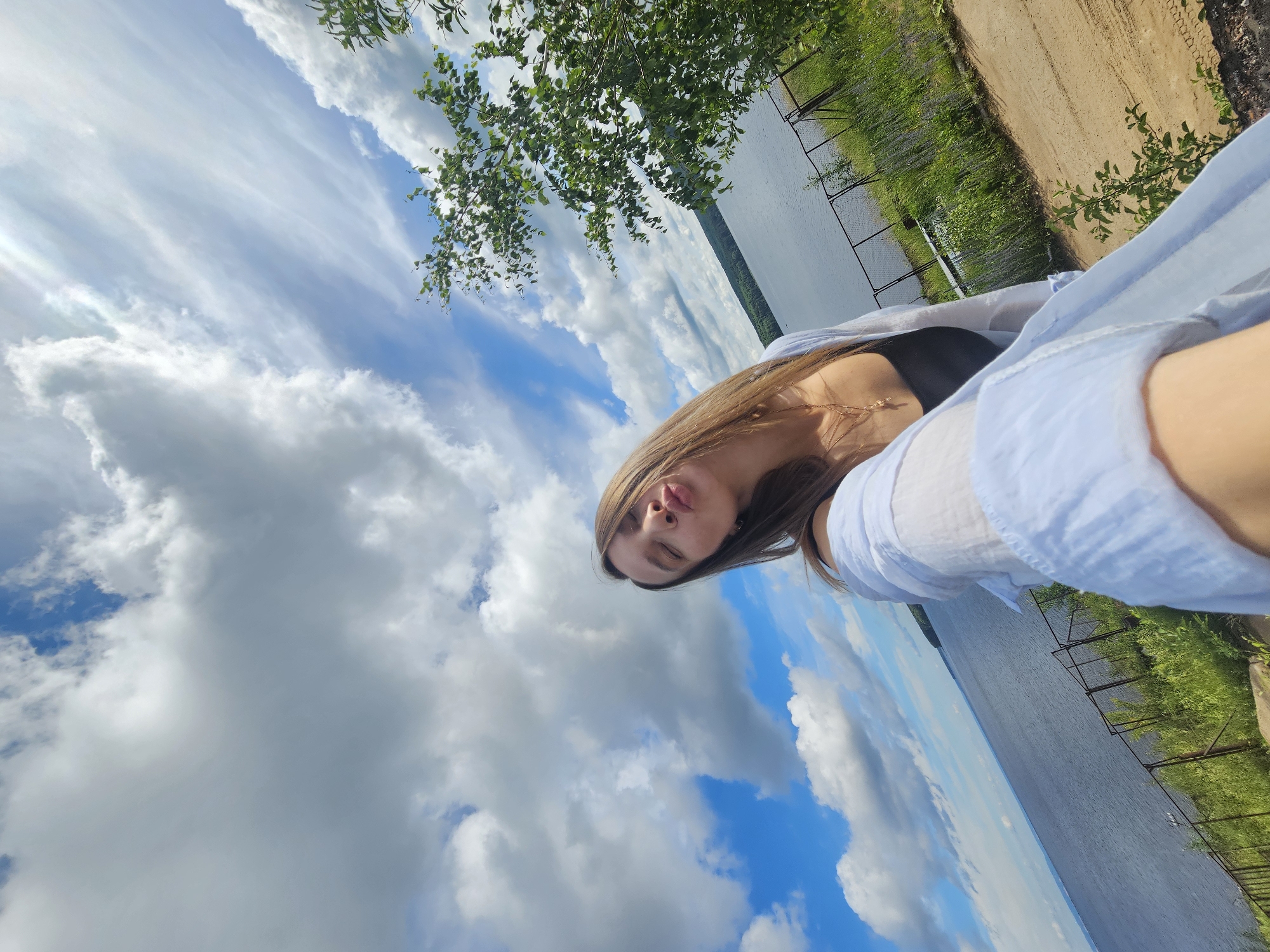 A woman is taking a selfie outdoors in Chaikovsky, Russia, with a body of water and a cloudy sky in the background. She is wearing a black top and a light blue open shirt. Her eyes are closed and her lips are pursed. A fence and vegetation are visible in the foreground, suggesting she is on a raised vantage point overlooking the water. The environment appears to be a park or natural area along the shore.