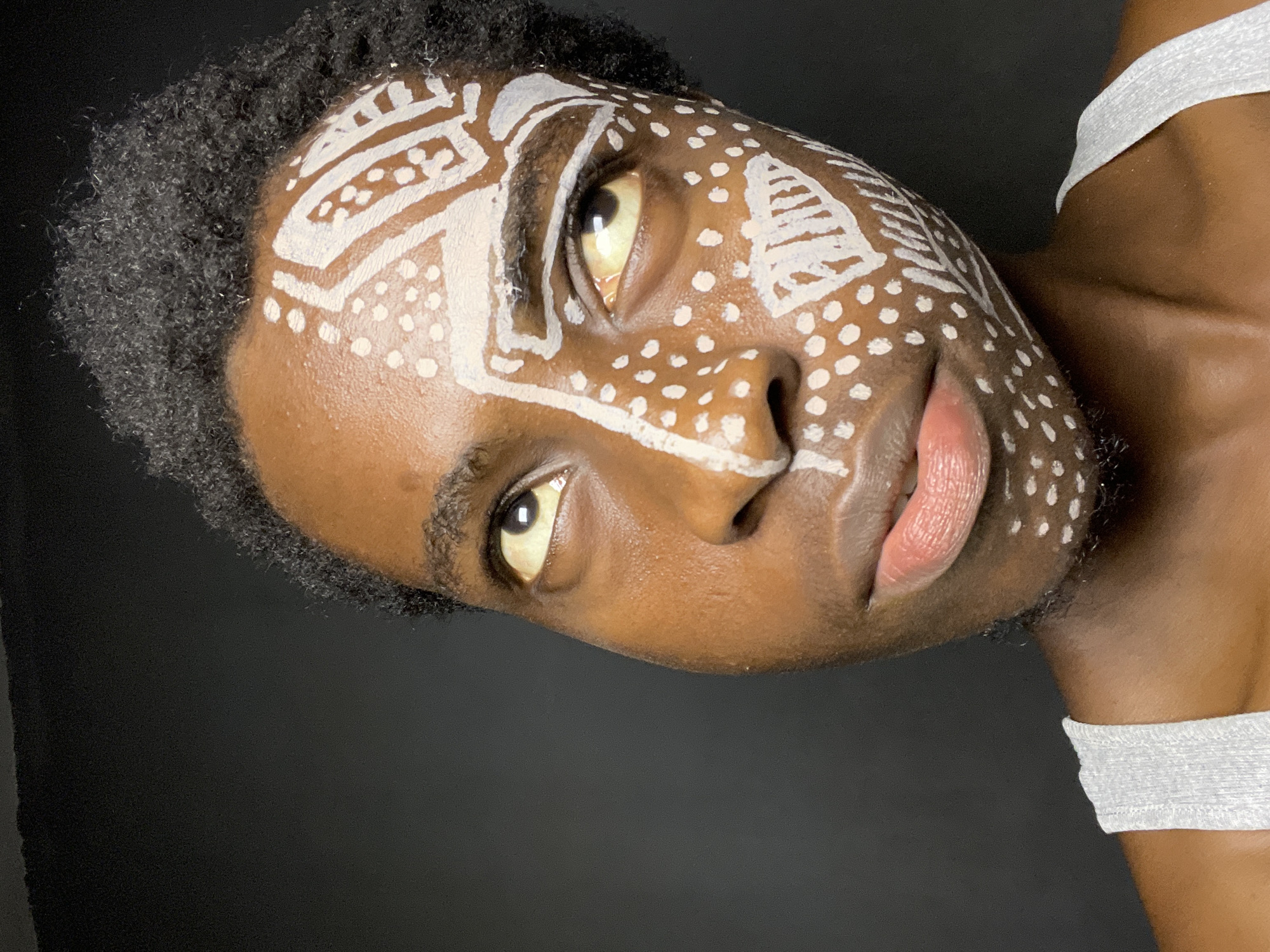 A close-up portrait shows a dark-skinned person with short, tightly curled black hair. Their face is adorned with white paint patterns, including geometric designs on the forehead and dotted patterns across the cheek and nose. The person's eyes are open, and their lips are slightly parted. They are wearing a white fabric strap over their shoulder. The background is a plain, dark gray or black color. There is no information available to confirm the location of this image is Nkpor, Nigeria.