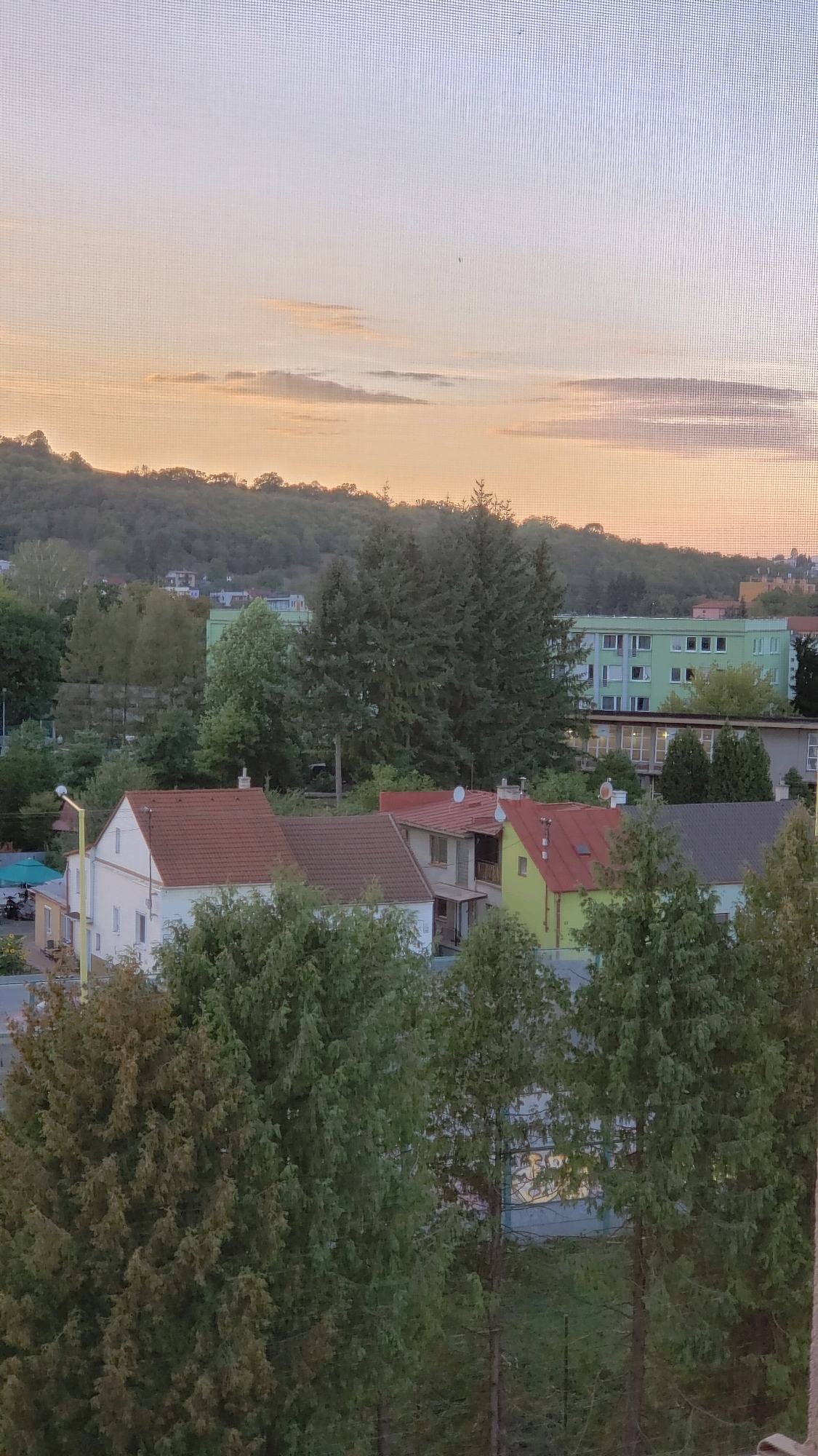 The image depicts a residential area in Prešov, Slovakia, viewed through a screen or mesh, possibly a window. The foreground is dominated by lush green trees. Behind the trees, several houses with red and gray tiled roofs are visible, some with white or colored walls. A larger, light green apartment building with numerous windows stands further back, set against a backdrop of a tree-covered hill. The sky above is a pale orange and blue gradient, suggesting either sunrise or sunset, with a few wispy clouds. The overall scene is one of a peaceful, urban-suburban landscape.