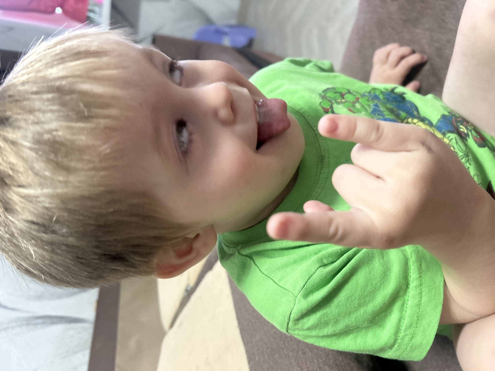 A young child is lying on a brown couch. The child has blond hair and is wearing a bright green t-shirt with an illustration on it. The child is sticking their tongue out and making a rock-on hand gesture with their right hand. The background is blurred, but a glimpse of a white object and a patterned wall can be seen. The location, Chaikovsky, Russia, is not verifiable from the image.