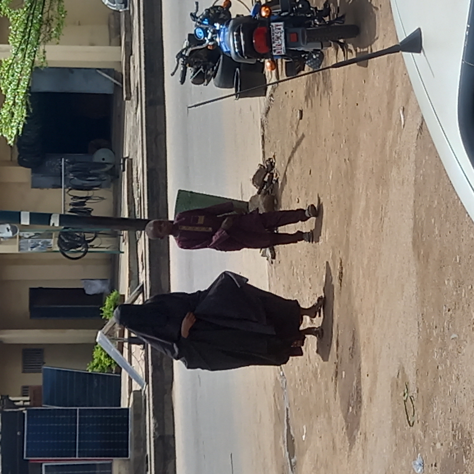 The image shows an urban street scene in Dadi Riba, Nigeria. A woman in a black niqab and high-heeled shoes walks on the unpaved roadside. Adjacent to her, a man in a maroon outfit, complete with dress shirt and pants, is also walking. A motorcycle is parked near the curb, and parts of a white vehicle are visible in the upper right corner of the frame. Solar panels are attached to the side of a building, and various items are stored under the building's open lower level.