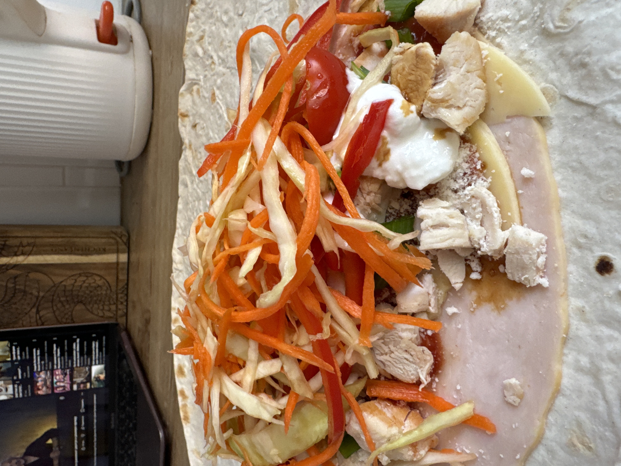 An overhead, slightly angled shot captures a partially assembled wrap or flatbread dish, laden with various ingredients, resting on a wooden surface.
The central focus is the food item, featuring a textured, light-colored flatbread that is open to reveal its abundant filling. Dominating the left side of the filling is a large pile of shredded orange carrots and lighter, possibly shredded cabbage or other leafy greens. Towards the right, chunks of cooked chicken are visible, along with slices of pale pink deli meat, and thin, light yellow slices of cheese. A dollop of white creamy sauce, likely yogurt, sour cream, or mayonnaise-based, sits atop the chicken, accented by a red cherry tomato half or sliced red pepper. Scattered green specks suggest chopped green onions or chives, and a fine white powdery substance, possibly grated cheese like Parmesan or feta, is sprinkled over parts of the filling.
In the upper left background, a white kitchen appliance with a small orange handle or button is partially visible. The entire food item rests on a wooden surface, possibly a cutting board or table. In the lower left corner, the edge of a laptop or tablet screen is visible, displaying a cluttered interface with numerous small, indistinguishable image thumbnails, suggesting a media library or streaming service. No legible text is discernible on the screen or elsewhere in the image. The scene appears to be set indoors, likely in a kitchen or dining area, with bright, even lighting.