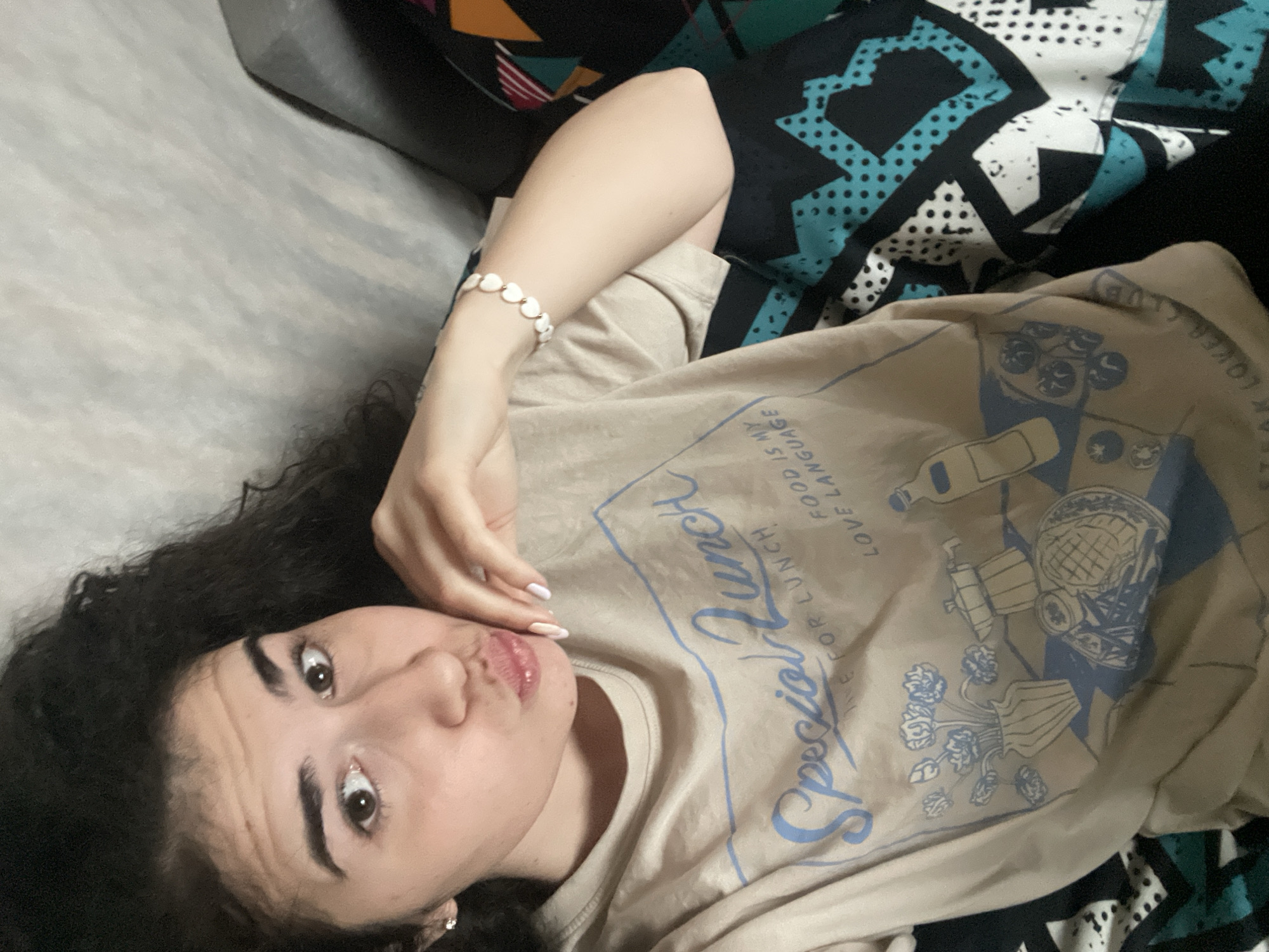 A young woman with curly dark hair is lying on a gray sofa and patterned cushions. She is wearing a beige T-shirt with a "Special Lunch" graphic in blue. Her left wrist is adorned with a white beaded bracelet, and she has light-colored nail polish. She is looking at the camera with a slightly pursed expression. The location is not discernible as Sofia, Bulgaria.