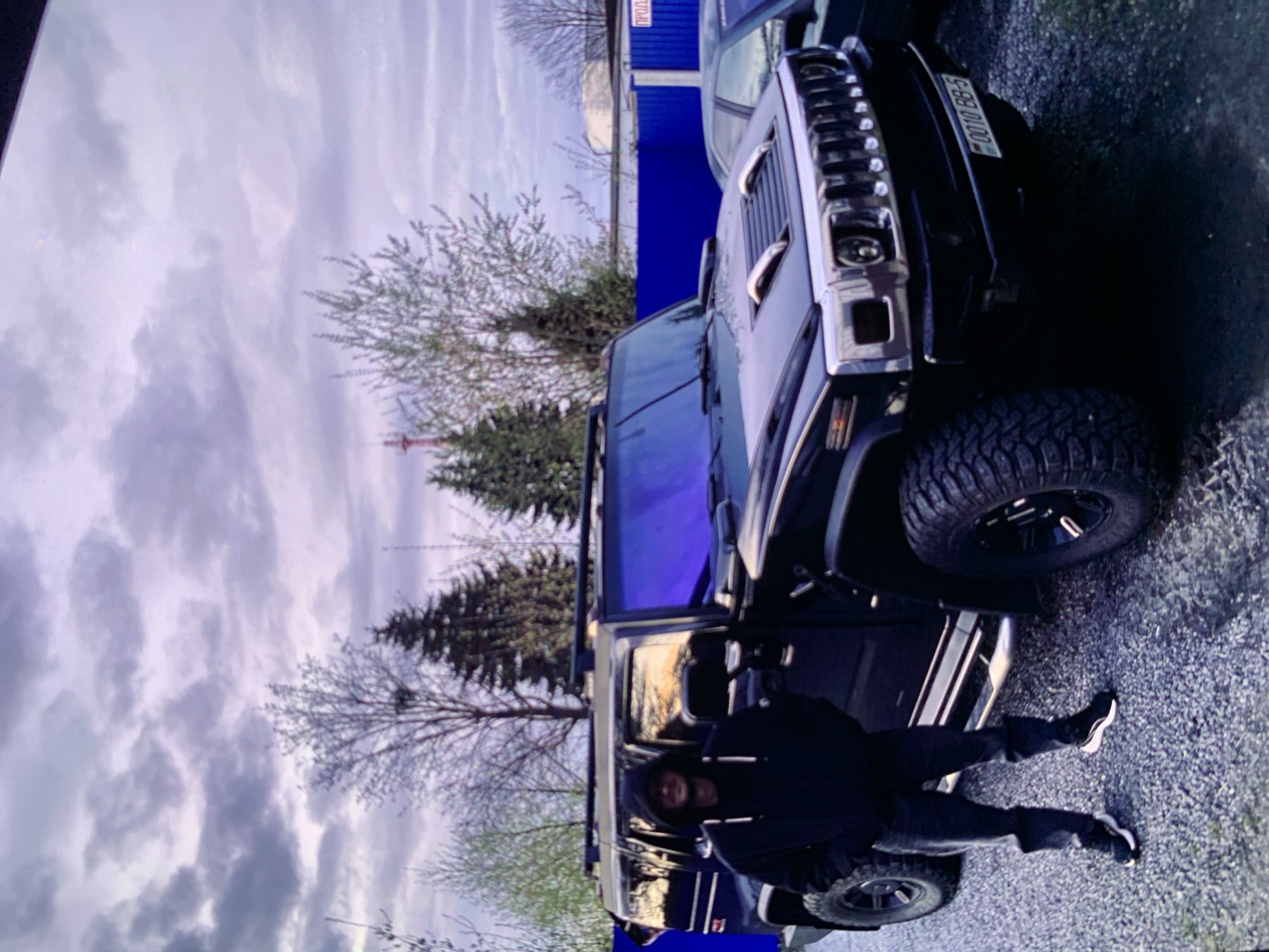 A person wearing a black hooded sweatshirt and dark pants stands next to a black Hummer H2. The Hummer has tinted windows and a license plate that reads "0010 BB-5". The scene appears to be outdoors in a gravel lot, possibly a parking area. In the background, there are trees, a blue fence, and a building. The sky is cloudy and has a purple hue, suggesting it might be dusk or dawn. The location is identified as Unknown, Unknown.