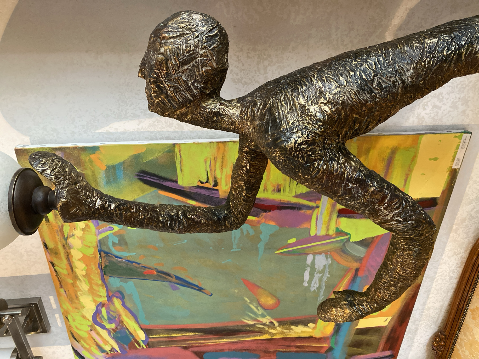 The image shows a bronze-colored statue of a person seemingly reaching forward. The statue is positioned in front of a large, colorful abstract painting. The background is a plain, textured wall. Part of a lamp is visible on the left. The location is unknown, and cannot be determined from the visual information available. No humans are present.