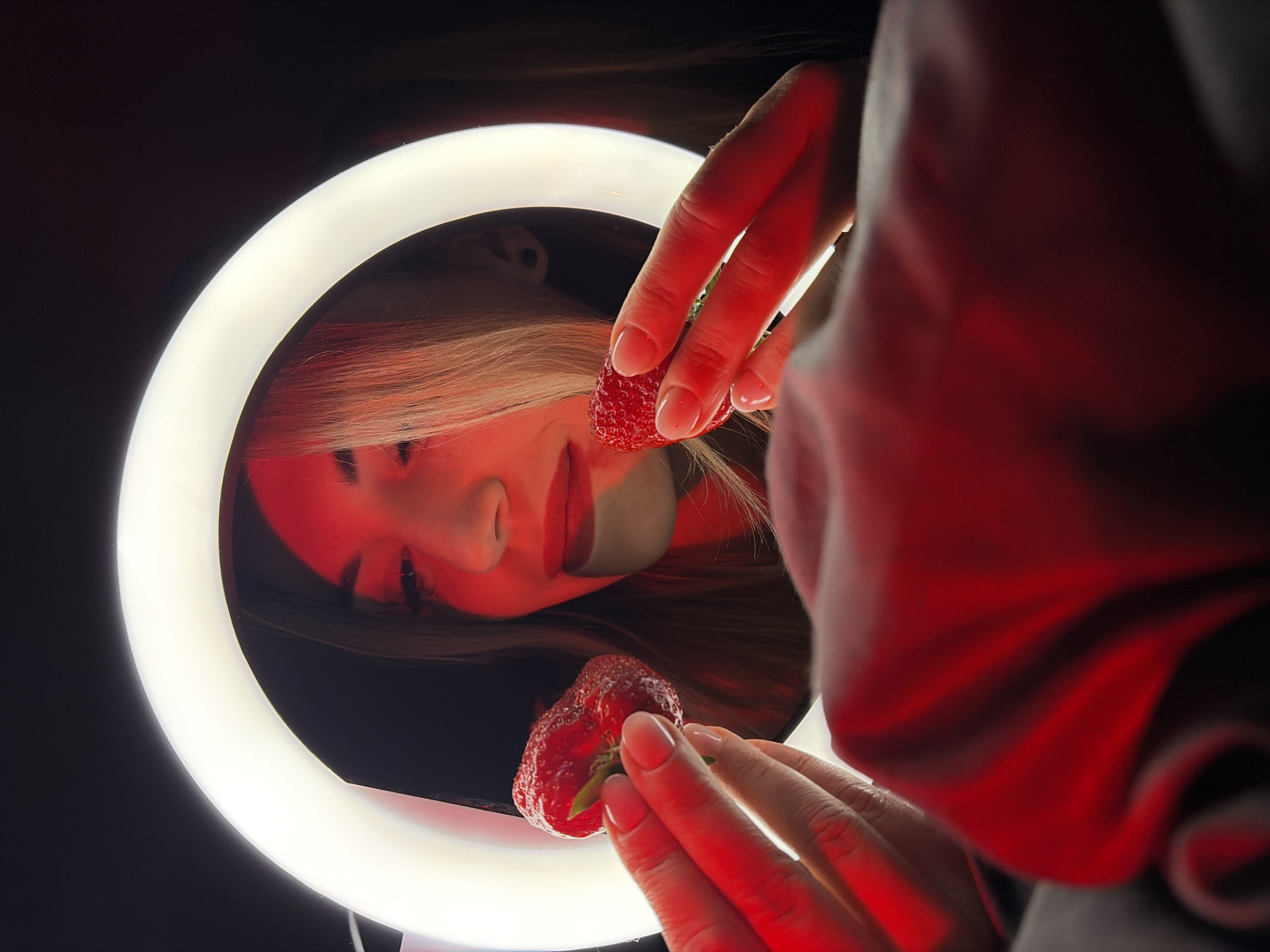 The image shows a woman's face reflected in a ring light. The woman has light hair and is wearing red lipstick. Two strawberries are held near the ring light by a hand with painted fingernails. The hand and part of a blurred sleeve are visible to the right of the ring light. The background is dark. There is no information about the location being in Budva, Montenegro.