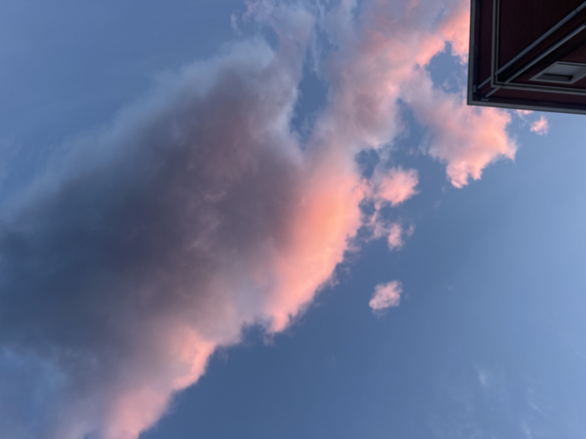 The image shows a view of the sky with clouds, taken from what appears to be a street-level perspective. The clouds are illuminated with a pinkish-orange hue, likely from the setting sun. The sky is a gradient of blue, transitioning from lighter near the horizon to deeper near the top of the image. The angle suggests that the camera is looking up. The roof of a building is visible in the top right corner of the frame. The image does not explicitly reveal the location in the city of Budva, Montenegro. The context provides no information about any people, subjects, objects, actions or interactions.