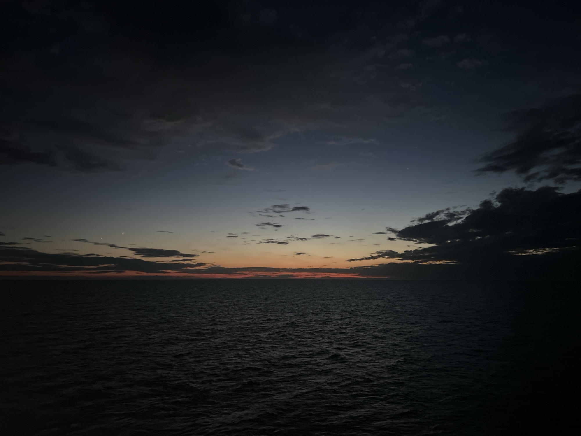 A wide-angle, high-angle outdoor nighttime shot shows a dark, choppy ocean under a deep blue and grey sky with scattered clouds. The horizon is dimly lit with a faint orange and pink glow from a setting or rising sun. Two small, bright celestial bodies are visible in the sky. The scene is dark and appears to be at sea.