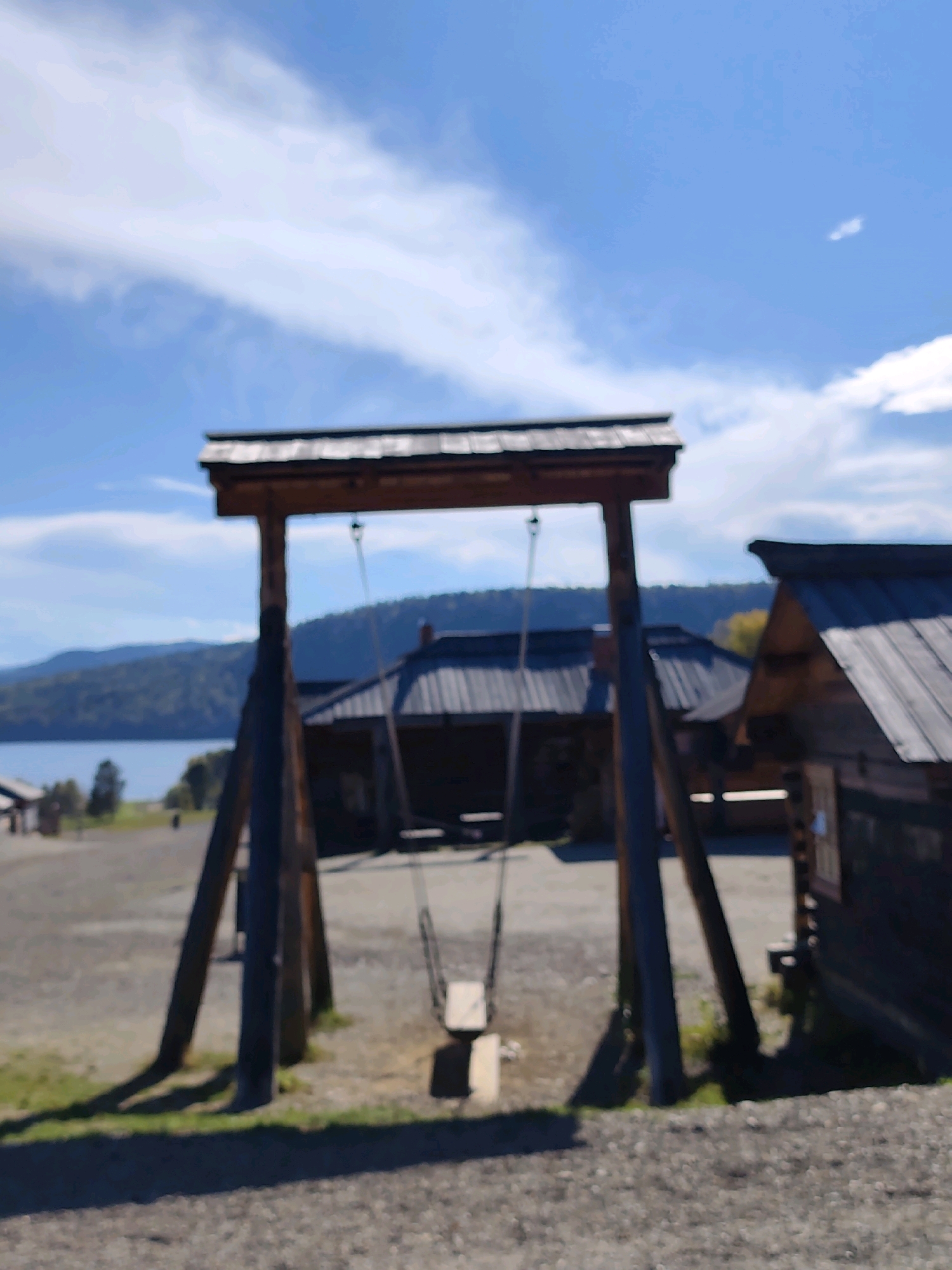 A wooden swing set stands in the foreground of an outdoor scene in the Museum of Wooden Architecture of the Taltsy, Irkutsk Oblast, Russia. The swing set is constructed with two large, angled wooden posts supporting a horizontal beam, from which a simple wooden swing is suspended by ropes. In the background, several wooden buildings with dark, shingled roofs are visible, characteristic of traditional Siberian architecture. A body of water and a tree-covered hill form the distant landscape under a blue sky with scattered white clouds. The ground is covered in gravel and patchy grass. There are no people visible in the image.