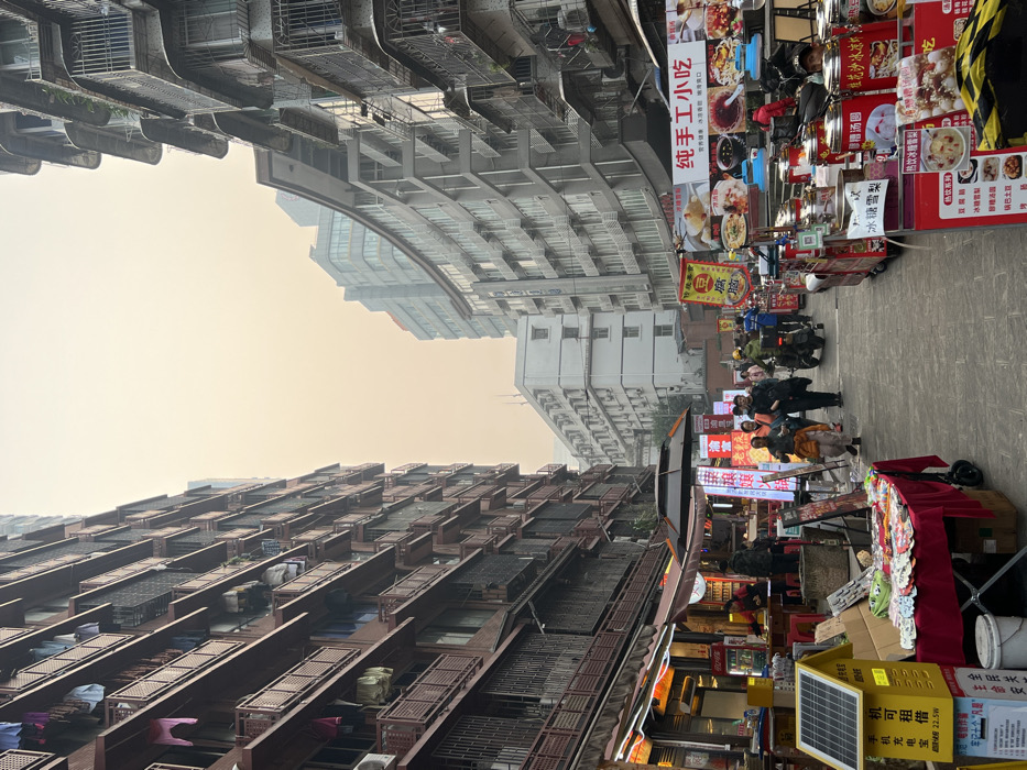 This image captures a narrow urban street in Yuzhong District, China, densely populated with buildings and commercial activity under a hazy, light orange sky.
On the left side, a tall, multi-story residential building with reddish-brown facades and numerous balconies extends upward. Clothes are visible drying on multiple balconies, and several external air conditioning units are affixed to the building. At street level, shops with brown awnings are present, one displaying a Coca-Cola logo. In the immediate foreground, a yellow box with a solar panel is labeled as a "mobile phone charging station" (手机充电站) with "22.5W" output. Adjacent to it, a table draped in a red cloth displays various small items for sale. Further down the left side, vendors operate under dark umbrellas, with red plastic chairs and tables indicating outdoor dining areas.
On the right side, a series of multi-story buildings primarily constructed of grey concrete features balconies, many secured with metal grilles. Further down the street, a more contemporary grey building with curved balconies and extensive glazing is visible, alongside a plainer white multi-story building. At street level, numerous mobile food stalls with red bases and silver food containers are positioned, displaying various snacks such as "冰糖雪梨" (rock sugar pear). Large banners with Chinese characters, including "纯手工小吃" (pure handmade snacks), and photographs of food items are attached to these stalls and buildings.
The paved street accommodates approximately 10-15 pedestrians, some walking away from the viewer. Two motorcycles or scooters are present, with one rider visible on a black scooter in the midground, suggesting active street traffic. A vendor is partially visible behind one of the food stalls on the right. The scene depicts a busy commercial thoroughfare within a densely built urban environment.