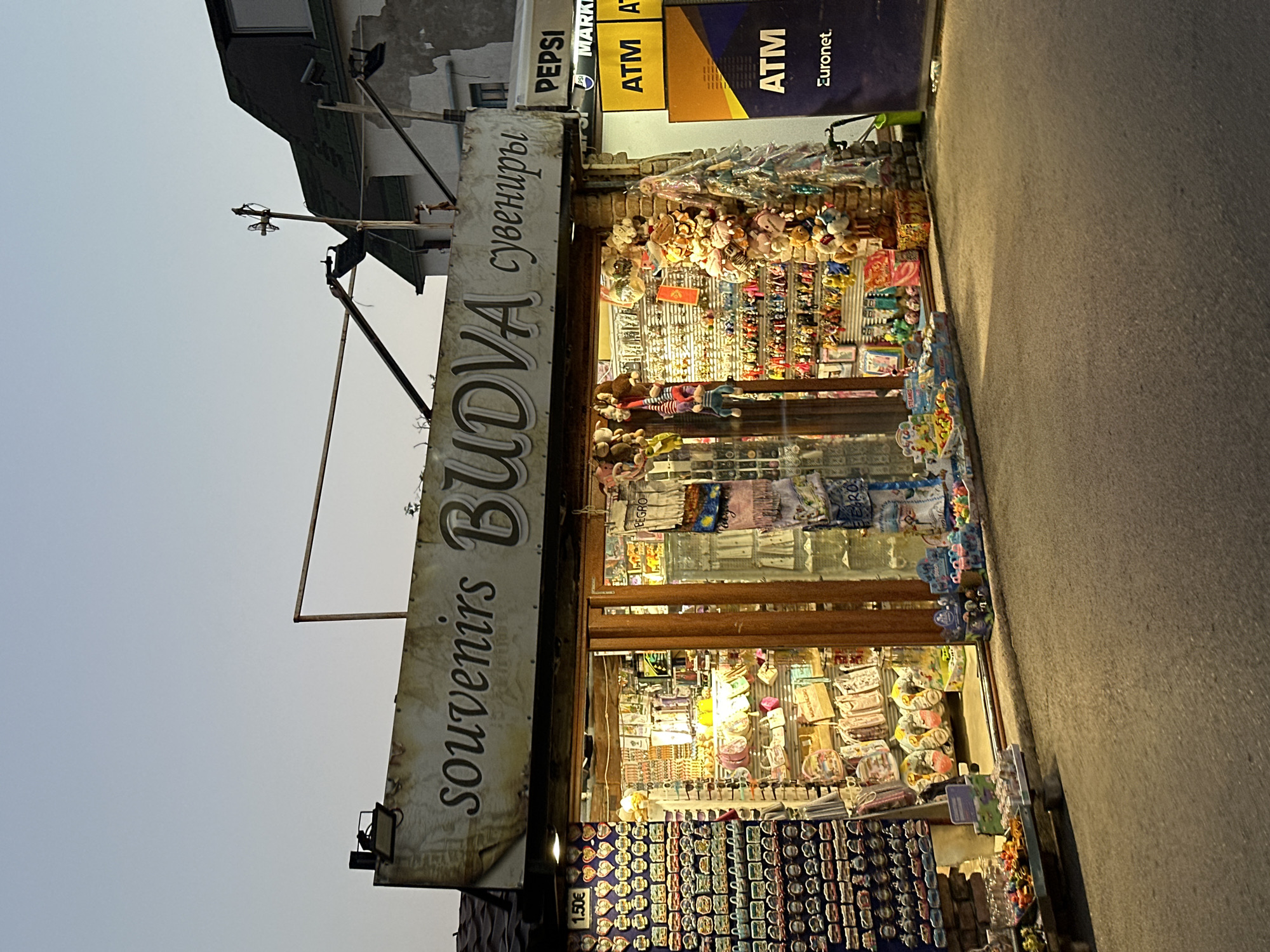 This is a frontal, eye-level, medium shot of a souvenir shop in Budva, Montenegro, at dusk. The shop is brightly lit from within, showcasing a variety of merchandise. The sign above the shop reads "souvenirs BUDVA" in English and "сувениры" in Cyrillic. To the right of the souvenir shop, there are two ATM signs, one yellow and one dark blue, both with "ATM" visible. A Pepsi sign is also visible above the ATMs.
The shop's facade features a large display window filled with shelves packed with items such as keychains, magnets, small bags, plush toys, and other trinkets. The exterior of the shop is constructed with brick on the right side. In front of the shop, on the pavement, are additional displays of merchandise, including small toys and candy. A wheeled shopping cart is parked to the right of the entrance. The street in front of the shop is paved and dimly lit, with shadows cast by the shop's lights. The sky above is a gradient of pale blue and grey, indicating twilight. No people are visible in the scene.