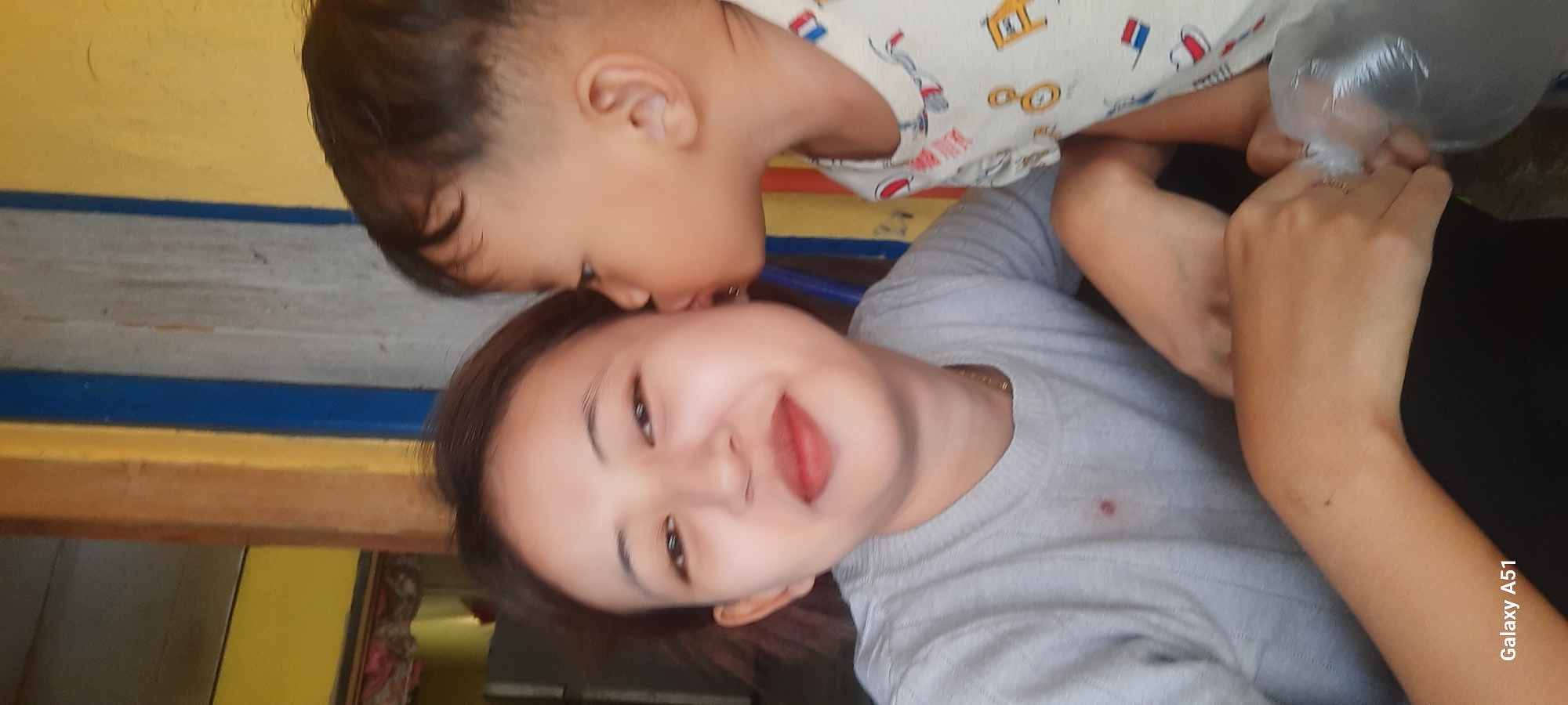 In Selopuro, Indonesia, a young woman with light brown hair and fair skin, wearing a grey t-shirt and a gold necklace, is photographed from the chest up. A young child, with dark hair and fair skin, is leaning in to kiss the woman's cheek. The background features a yellow wall, a brown wooden post, and a blue stripe. The woman is smiling slightly, with red lipstick on her lips. The child is wearing a white t-shirt with colorful illustrations. The lower right corner of the image displays the text "Galaxy A51".