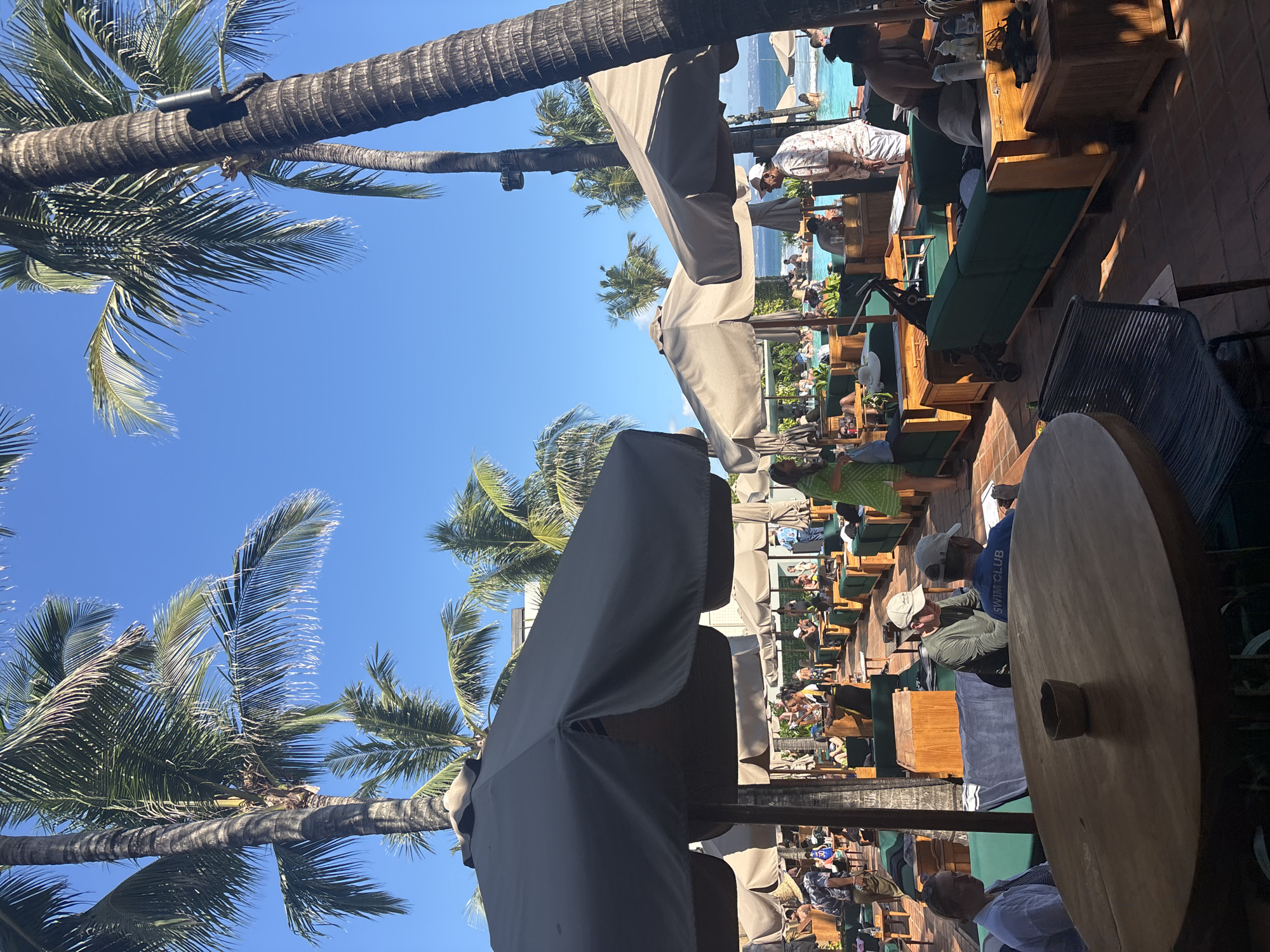In North Kuta, Indonesia, a bustling outdoor poolside lounge is captured under a clear blue sky, with towering palm trees framing the scene. Patrons are seated on dark green lounge sofas arranged in rows, some under large, neutral-colored umbrellas, while others bask in the sunlight. Several individuals are in the turquoise swimming pool in the background. A wooden table with a small bowl is prominent in the foreground, with a dark woven chair beside it. The overall atmosphere suggests a lively resort environment, likely during the daytime.