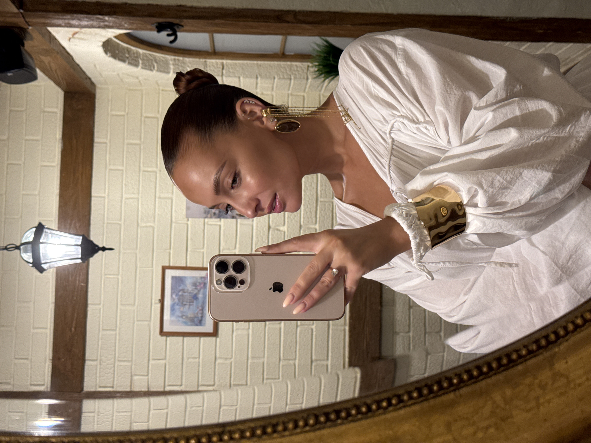 A woman is taking a selfie in a mirror. She is wearing a white, ruffled blouse and large gold earrings. She has her hair pulled back in a bun. The background appears to be a bathroom with brick walls and a wooden beam. A framed picture hangs on the wall, and a lamp is visible.