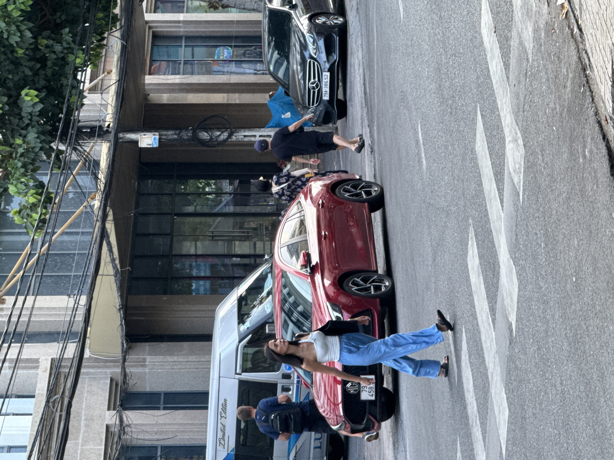 This is a street scene in Nha Trang Ward, Vietnam, featuring a woman crossing a pedestrian crosswalk. She is wearing a white strapless top and loose blue pants, carrying a dark bag over her left shoulder. To her left, a red sedan is stopped, and behind it, a white van with "Limited Edition" printed on its side is visible. To the right of the woman, a man in a black t-shirt and shorts is walking away from the camera, and further right, a dark sedan with a Mercedes-Benz emblem is parked on the street. The background shows a building with multiple windows and an overhead arrangement of electrical wires.