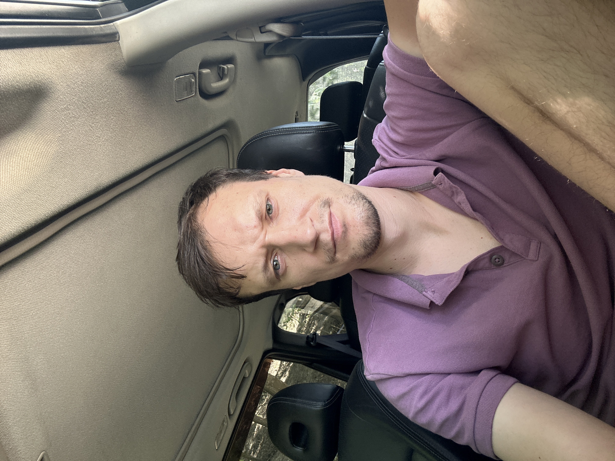 A man with short brown hair and a light beard is sitting in the driver's seat of a car. He is wearing a purple polo shirt. The car has black leather seats and a sunroof. The background visible through the windows appears to be a wooded area. The image does not provide any information about the location in Makiv, Ukraine.