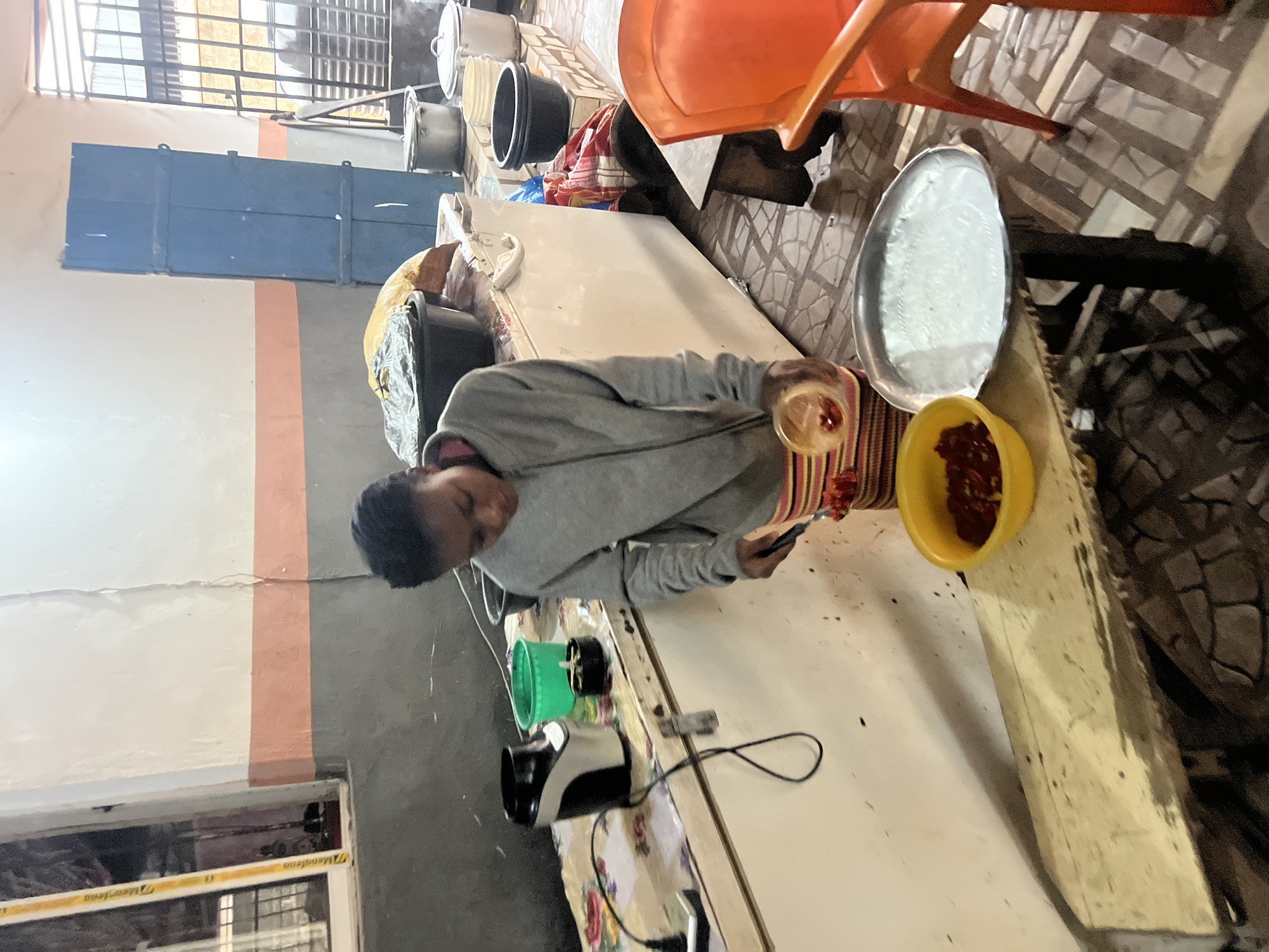 In Nkpor, Nigeria, a young person is seen preparing food in what appears to be a kitchen or food preparation area. The individual, wearing a gray hooded sweatshirt and striped pants, is focused on a bowl of red food, possibly tomatoes or peppers, as they transfer it with a utensil into a smaller container. Next to the bowls, a silver tray with a white substance, perhaps a dough or paste, sits on a wooden stool. In the background, a white refrigerator or freezer is visible, along with various cooking utensils and containers, including a black bowl containing what looks like food covered with foil. The room has tiled floors and white and pink walls, with a blue door and a window visible in the background. The overall scene depicts a common domestic or commercial food preparation setting.