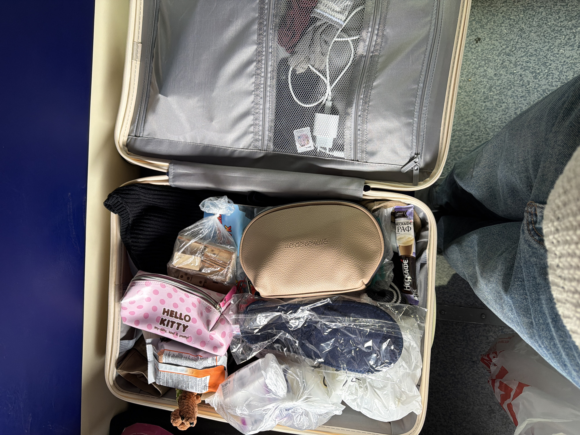 The image shows an open suitcase packed with various items. In the top half of the suitcase, which is open, there is a mesh pocket containing electronic chargers and cables. The bottom half of the suitcase is filled with clothes, bags, and personal items. Some visible items include a black knitted garment, a bag of what appears to be biscuits, a beige makeup bag labeled "tre de Beauté," a Hello Kitty pencil case, snacks, and a pair of blue denim slippers in a plastic bag. The bottom portion of the suitcase also contains small travel-sized items and a small stuffed animal. Part of a person's leg, clad in denim jeans, is visible to the right of the suitcase. The photo was taken in the city of Зелёный Дол, Russia.
