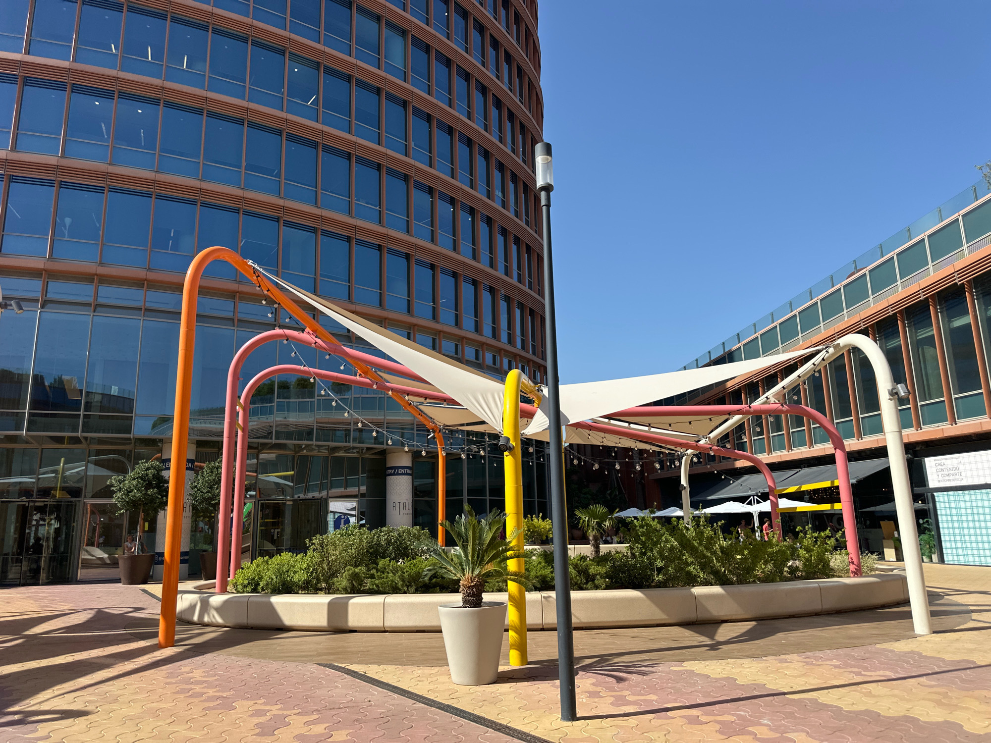 The image captures an outdoor scene in Seville, Spain, under a clear blue sky. In the foreground, a multi-colored paved ground is visible, with yellow, orange, and red interlocking tiles casting long shadows from overhead structures. A modern, curved building with a facade of glass windows and brown framing dominates the background. In front of this building, a shaded area is created by a tensile fabric canopy supported by colorful metal poles in orange, pink, and yellow. String lights are draped across the canopy.
Several planters with green foliage and a small palm tree in a light grey pot are situated within the shaded area. A black lamppost stands to the right of the frame. In the middle ground, through the entrance of the building, a sign with the text "ATALI" is partially visible. Further back, another building with a similar glass and brown framing design is visible, with an awning and outdoor seating areas. A few people are faintly visible in the distance, appearing to be patrons of the establishment. The overall scene suggests a contemporary urban plaza or commercial space designed for outdoor gathering.