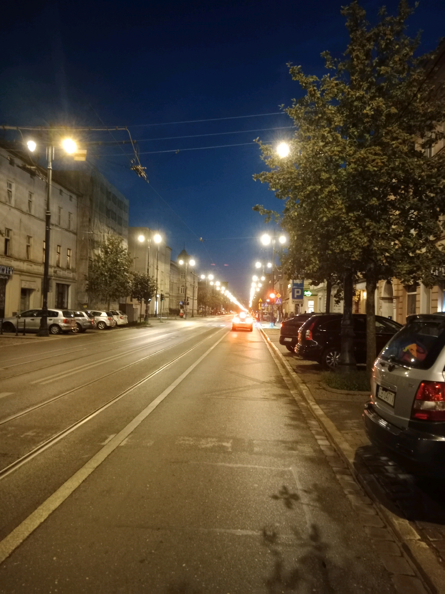 The image depicts a street scene in Bydgoszcz, Poland, at dusk. The perspective is from a sidewalk, looking down a street lined with buildings and trees. Streetlights illuminate the road, which has tram tracks running through it. Several cars are parked along the sidewalk and driving on the street. One car in the foreground has a sticker on its rear window. The sky is a deep blue, indicating twilight.