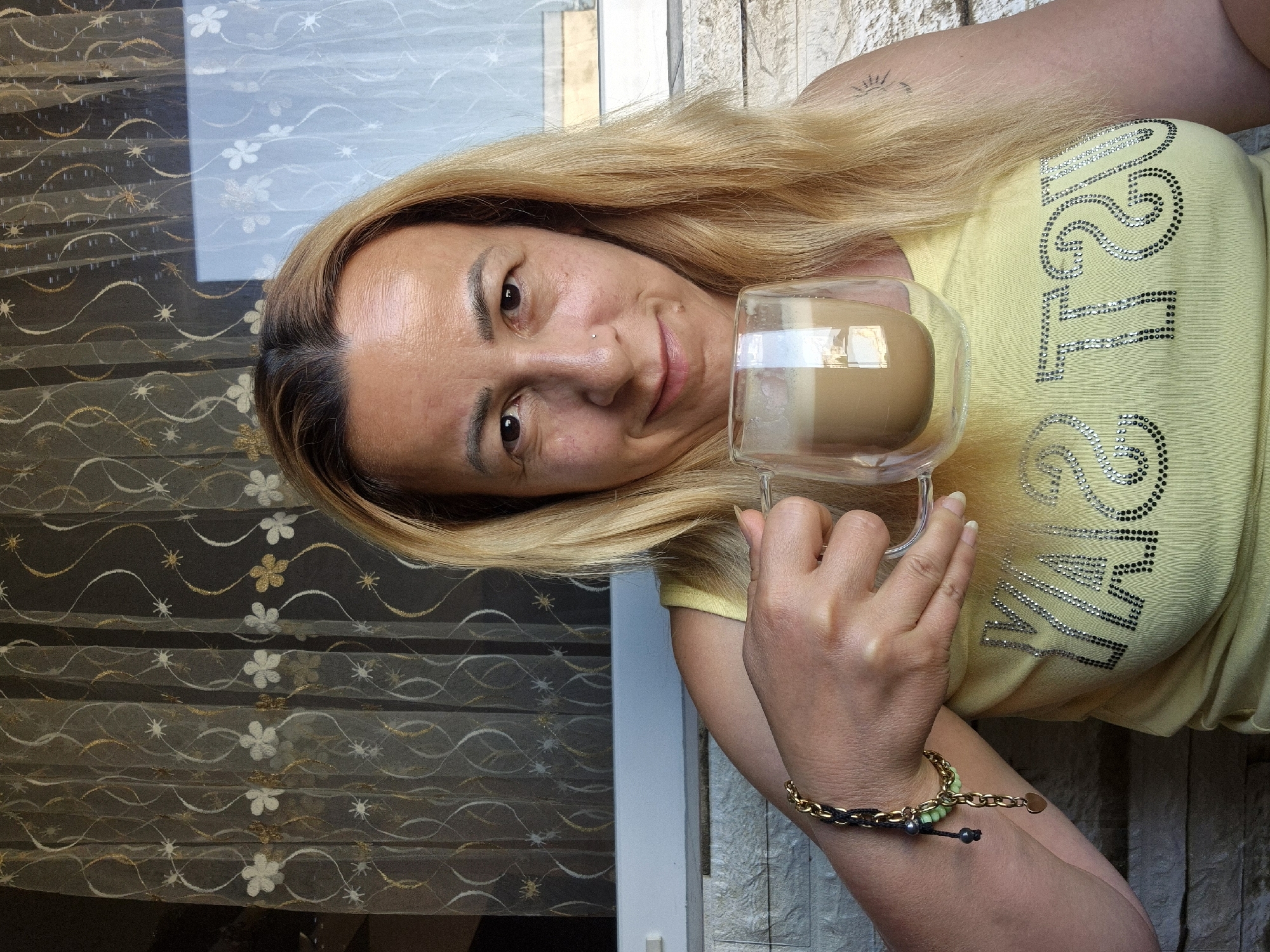 A woman with blonde hair and light-colored skin is holding a glass mug filled with a light brown beverage in her left hand. She is wearing a yellow t-shirt that appears to have a word or phrase printed on it in silver, sparkling letters. Her right arm is adorned with a bracelet made of black and gold links, with several green beads and a small gold heart charm. The woman is looking directly at the camera with a slight smile. Behind her, a patterned curtain with white and gold floral designs is visible, along with what appears to be a window with a light blue tint. The background also shows a stone-like wall texture. The scene is illuminated by natural light. The image seems to be a selfie.