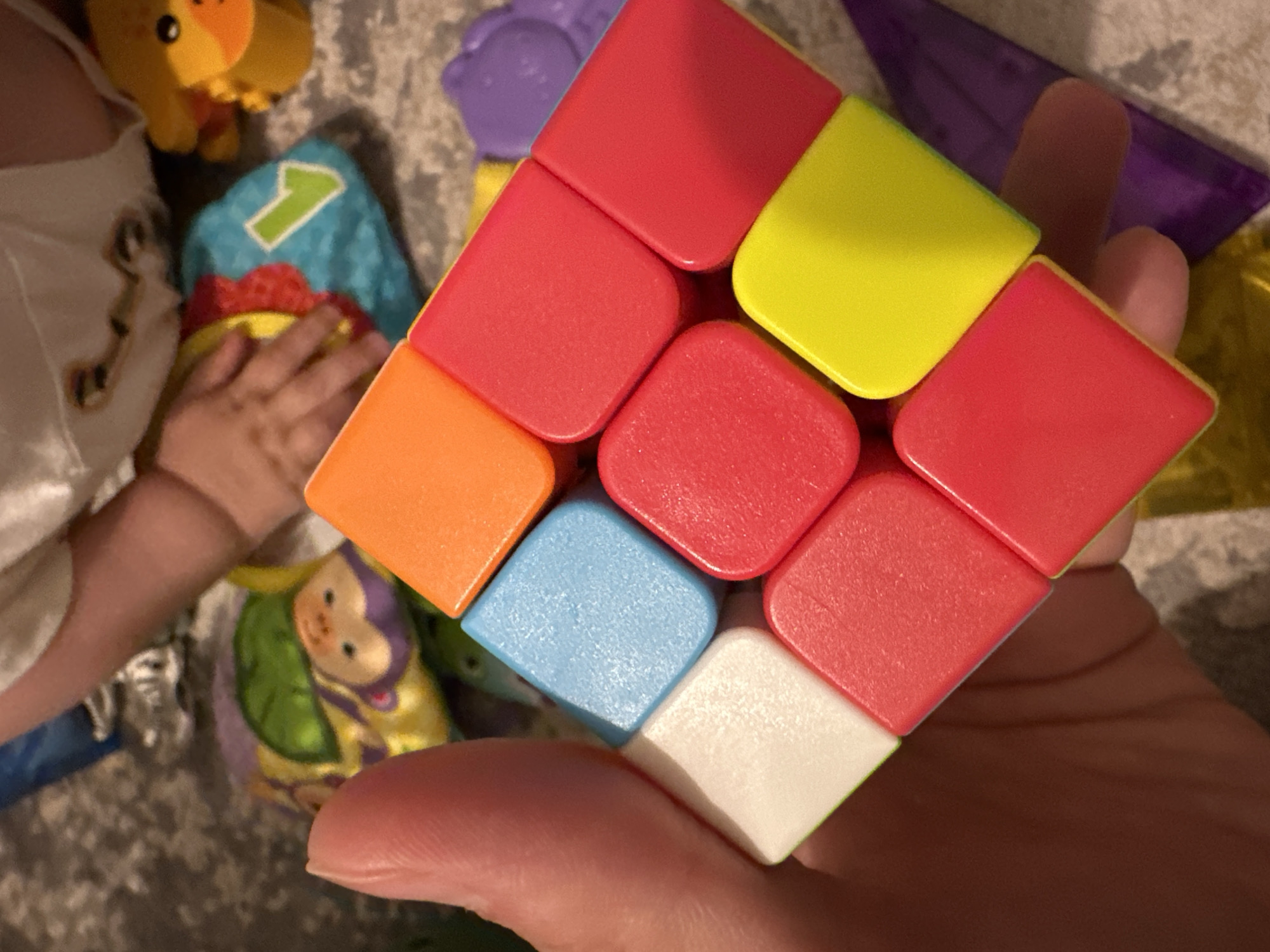 A hand holds a Rubik's Cube with red, yellow, orange, blue, and white faces visible. A child's hand rests near a plush toy featuring a numeral "1." Other toys, including a purple piece and a toy with a monkey face, are scattered in the background. The setting appears to be indoors. No specific location in Fethiye, Turkey, is discernible from the image.