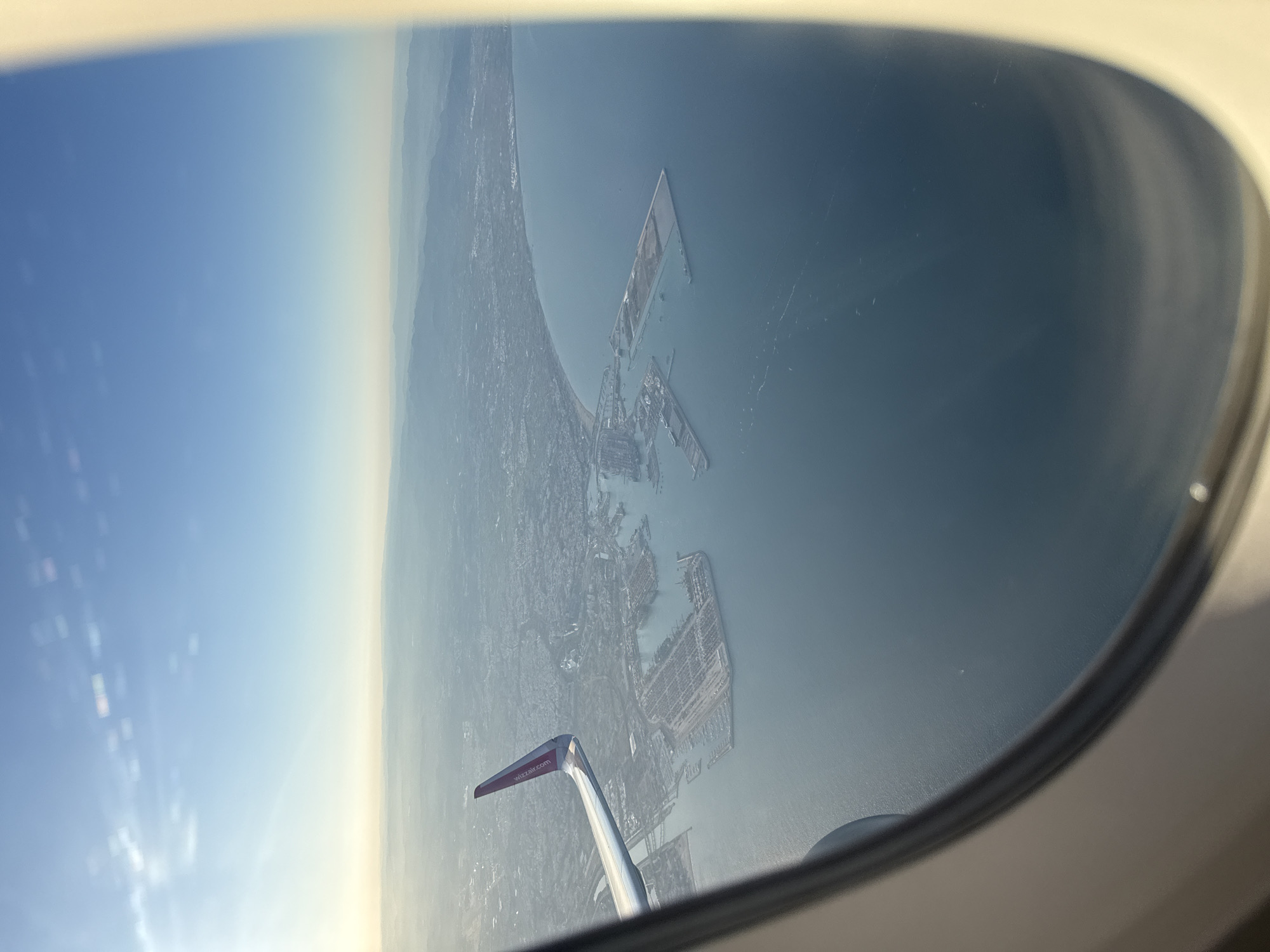The image is taken from inside an airplane looking out the window. The view is a city and coastline, and a large body of water. The wing of the airplane is visible in the bottom center. Based on the wing colors (white, purple, red) and lettering of wizzair.com, the image is from a Wizz Air flight. The city is likely Sofia, Bulgaria as the aircraft is Wizz Air, which has a base in Sofia, and the landmass features are consistent with the Bulgarian Black Sea coastline.