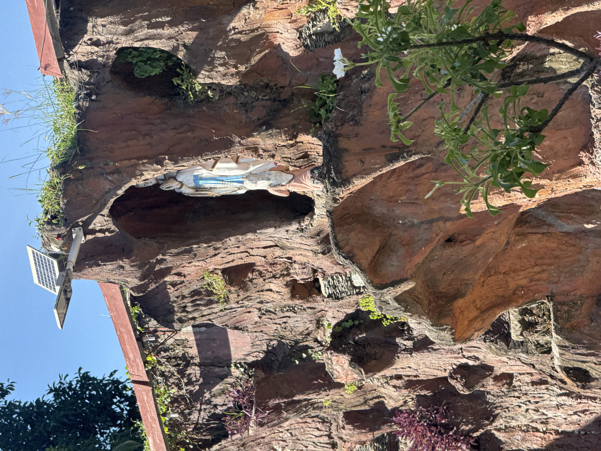 The image shows a statue of the Virgin Mary nestled within a man-made grotto that resembles a rock formation. The grotto is part of a larger religious structure, possibly a church or a shrine, located outdoors. A solar-powered light fixture is mounted on top of the grotto structure. Lush green vegetation and some purple plants are growing around the grotto, adding a natural element to the scene. The sky is clear and blue, suggesting a sunny day. The statue itself depicts the Virgin Mary in prayer, dressed in white and blue robes. The overall impression is one of religious devotion and natural beauty. This religious site is located in Davao City, Philippines, as indicated by the presence of typical Filipino religious imagery and landscaping.