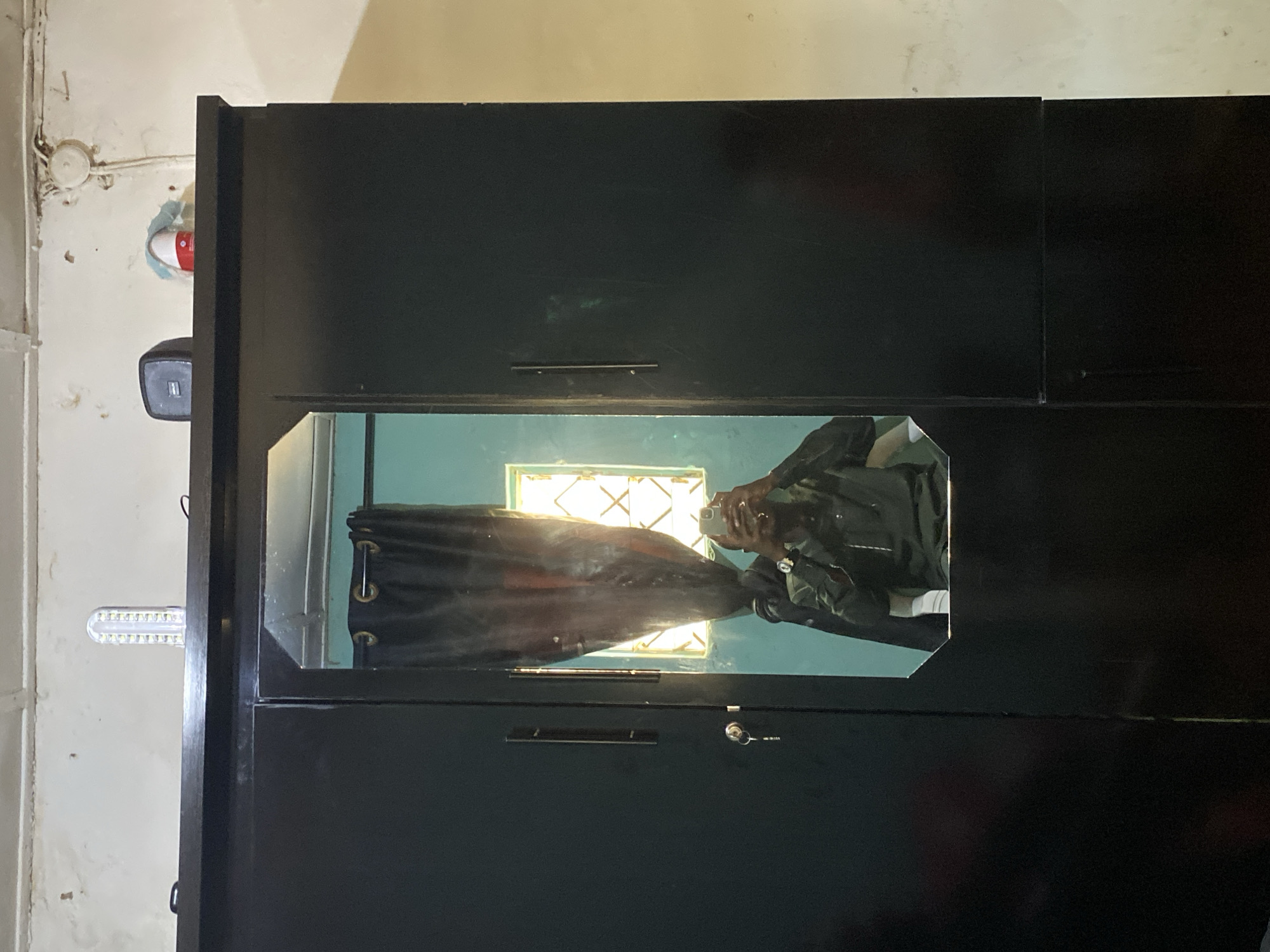 The image shows a black wardrobe or cabinet, likely indoors against a light-colored wall. The wardrobe features a mirrored section where the reflection of a person taking a photo with their smartphone is visible. A curtain is also reflected, along with a window behind the person, suggesting an interior scene. The light fixture is possibly led and located to the side of the wardrobe. Signs of wear and some staining appear on the wall next to the wardrobe. It is not possible to determine the location in Bukuru, Nigeria, from the given image.