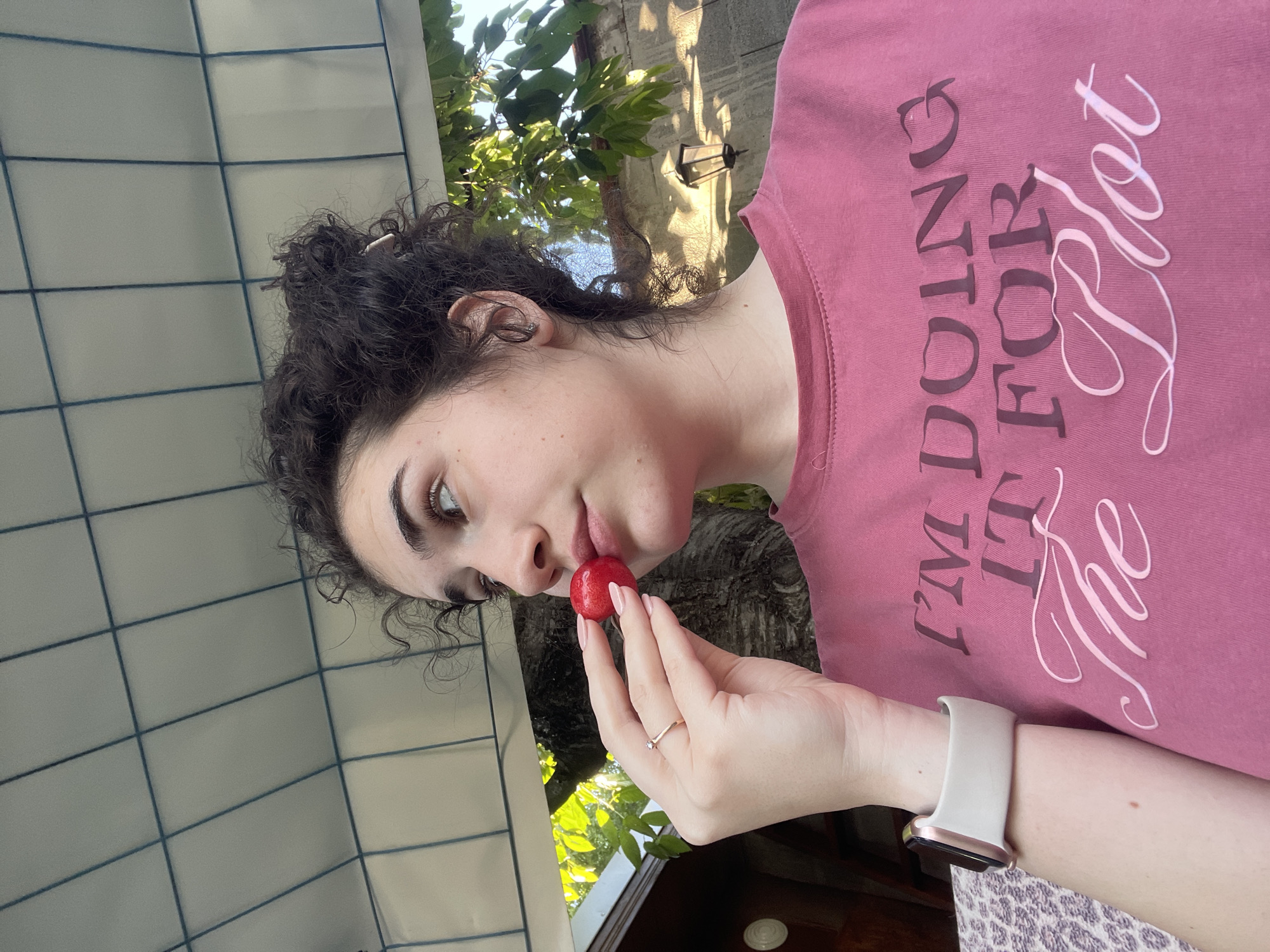 A woman with dark curly hair holds a red fruit near her mouth, possibly about to eat it. She wears a pink shirt with the text "I'm doing it for the plot" and a light-colored smart watch on her left wrist. She also has a ring on her left hand's ring finger. The background includes a tiled wall, foliage, and what appears to be a building or structure, possibly outdoors. Location details are not discernible to confirm Sofia, Bulgaria.