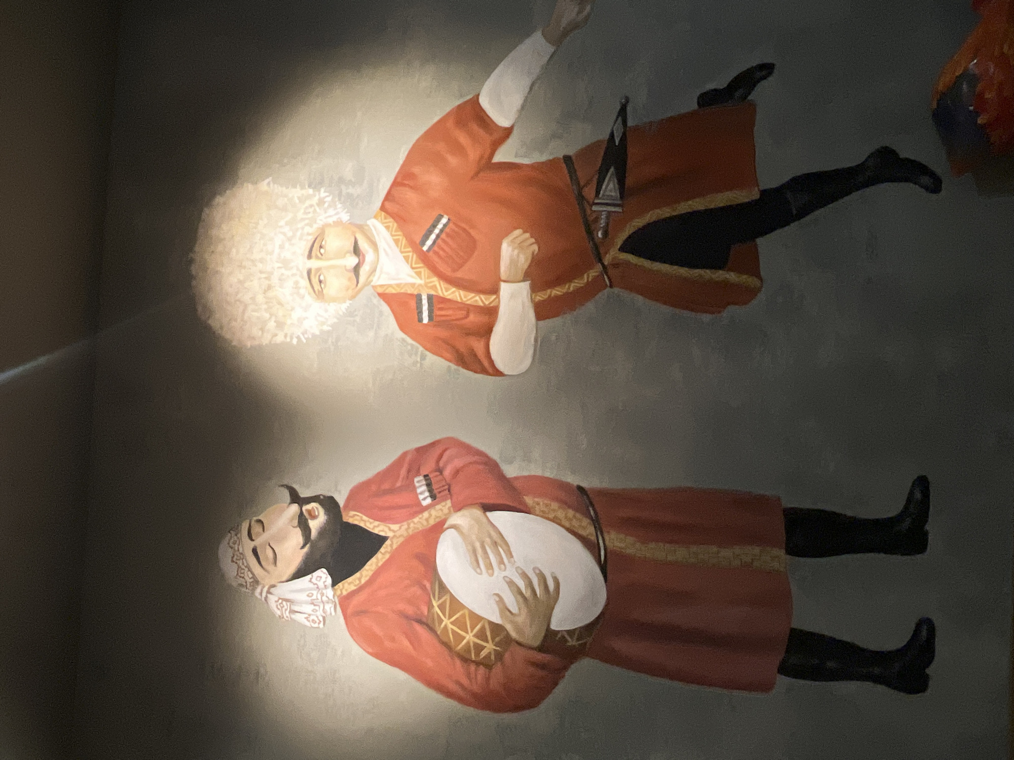 The painting depicts two men in traditional attire, possibly from the Caucasus region, set against a dimly lit, textured background. The man on the left wears a red robe, a white and patterned head covering, and holds a large drum. He has a dark beard and mustache and appears to be singing or chanting with his eyes closed. The man on the right is dressed in a similar red robe with a white collar and a large, fluffy white hat. He is in a dynamic pose, suggesting movement, and has a mustache. A dagger is visible tucked into his belt. The lighting highlights the figures, creating a dramatic effect. The style is illustrative, with bold colors and simplified forms. The painting is located in Khimki, Russia.