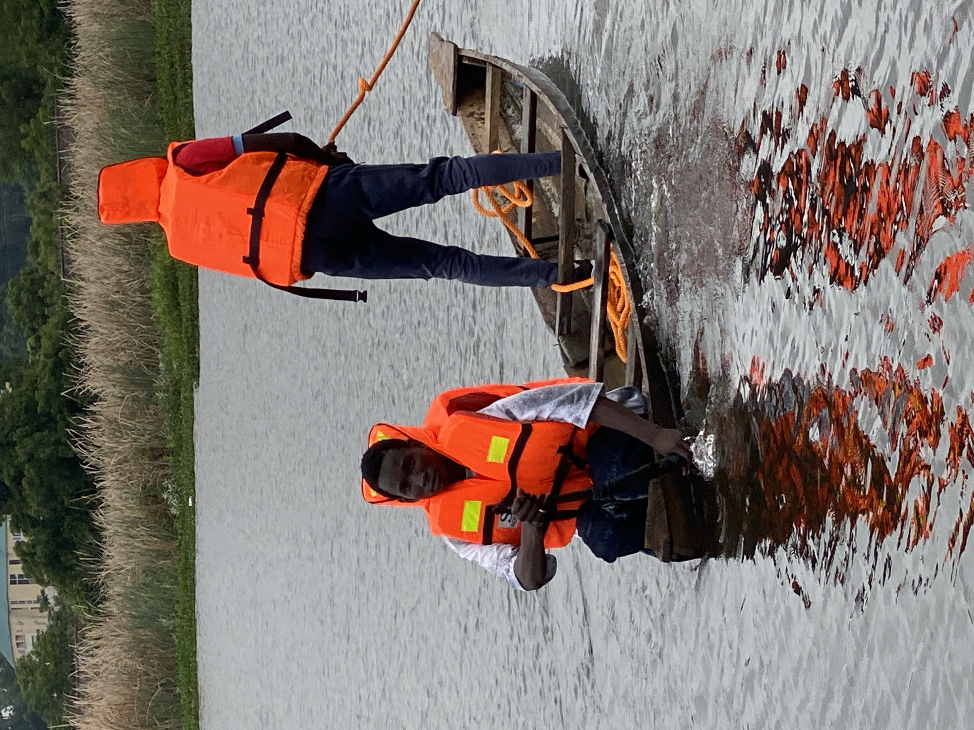 In Kawo, Nigeria, two individuals are in a wooden boat on a body of water. The person in the foreground is kneeling and appears to be operating the boat with a paddle. They are wearing an orange life jacket over a white shirt and dark pants, and they are looking towards the camera. Behind them, a second person stands in the boat, also wearing an orange life jacket over a red shirt and dark pants. This individual is holding an orange rope, which extends out of the frame. The boat is partially submerged in the water, and orange ropes are coiled inside. The environment includes dense vegetation and buildings in the background, suggesting an urban or semi-urban setting. The water is calm, with reflections of the boat and life jackets visible on the surface.