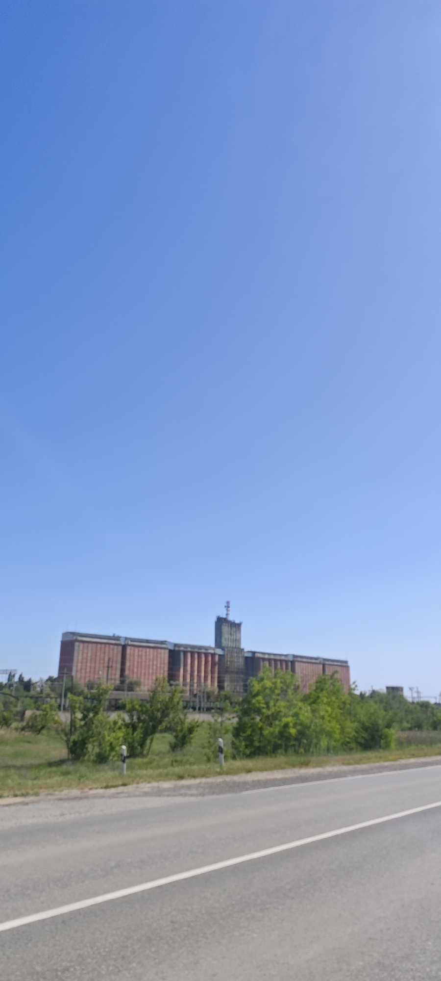 The image shows a long, horizontally-oriented building in Dubovskoye, Russia. The building has a central tower structure with a possible antenna on top. The main portion of the building features multiple sections, predominantly red, with gray accents. A line of green trees and bushes stands between the building and a gray asphalt road with a white line marking. The sky is a clear, bright blue.