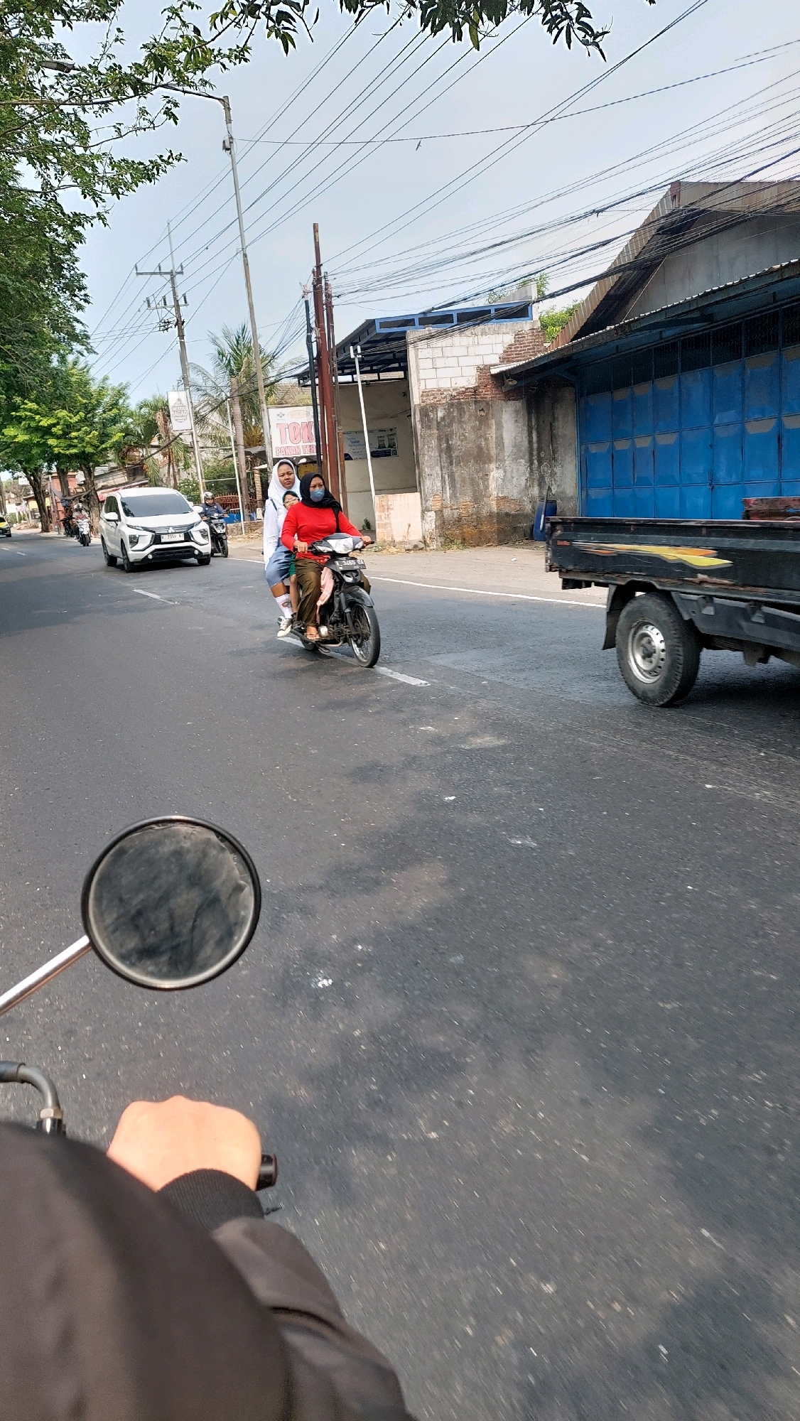 The image depicts a street scene in Ketimang, Indonesia. In the foreground, a person's hand and arm, wearing a black jacket, are visible, holding onto the handlebars of a motorcycle, as indicated by a rearview mirror. On the road, a motorcycle is carrying three people: a driver wearing a red hijab, a person in the middle wearing a white hijab, and a child behind them. They are traveling on the asphalt road. To the left, a white car is passing by, with a motorcycle behind it. Further down the street, a black flatbed truck is parked on the right side of the road, with a blue industrial-style door visible on a building behind it. The environment appears to be a commercial or industrial area, with buildings, utility poles, and overhead wires present. Trees with green foliage are visible on the left side of the street. The sky is clear and bright.