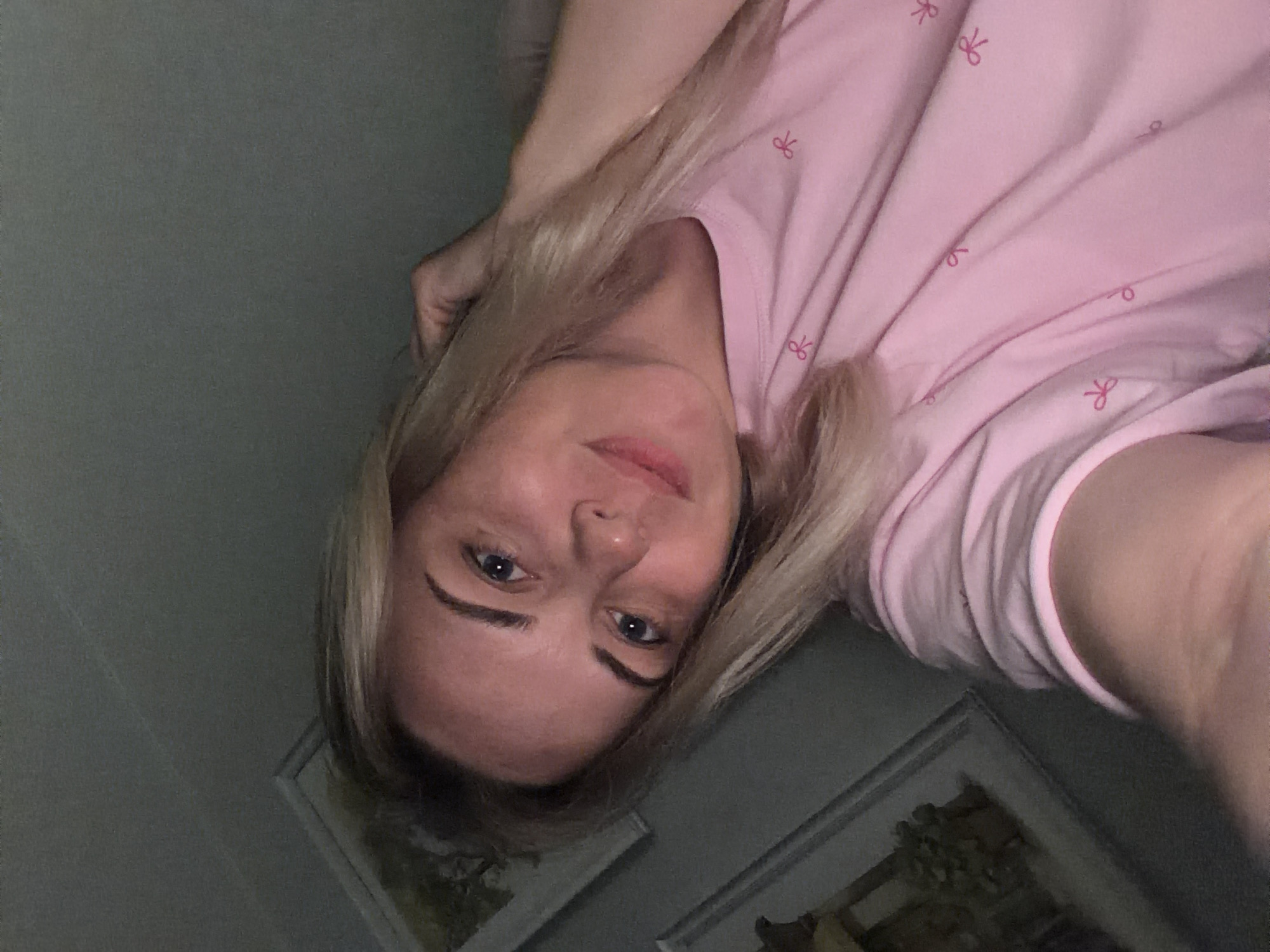 A close-up, head-and-shoulders selfie features a young woman with blonde hair and blue eyes. She is wearing a pink t-shirt with a pattern of small pink bows. Her head is tilted to the left, and her right hand is raised to her hair. She is looking directly at the camera with a slight smile. The background is dimly lit and appears to be indoors, with a framed picture visible on the left. The overall impression is a casual and relaxed pose. There is no identifiable information in the image that would indicate its location in Votkinsk, Russia.