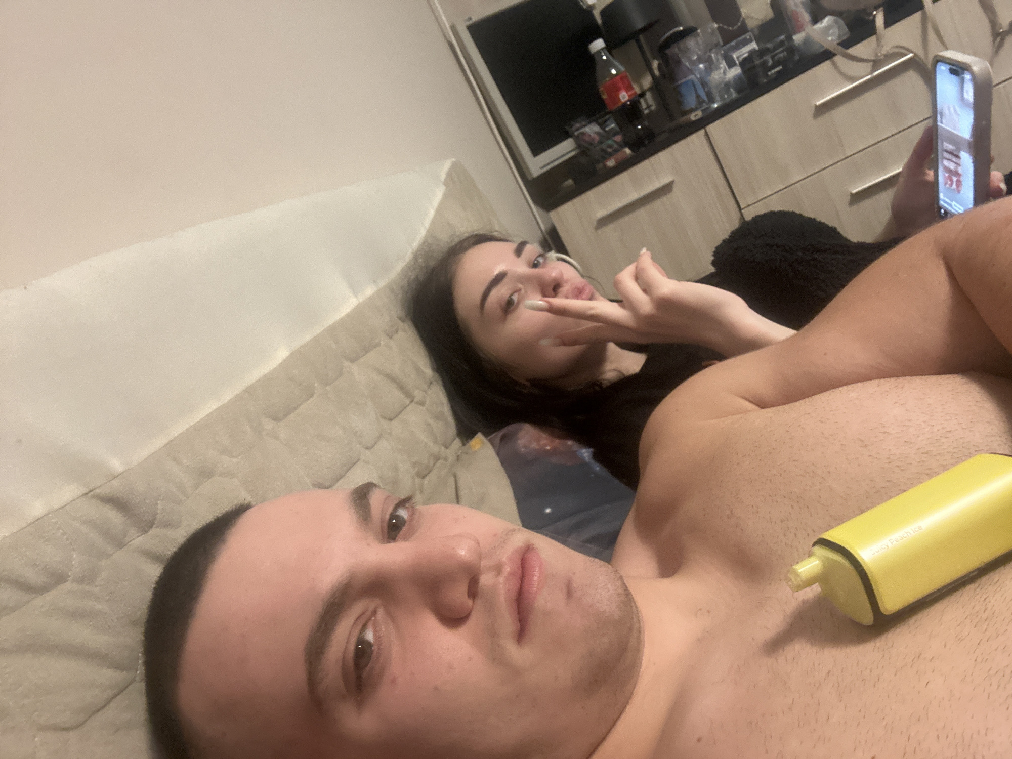 **Scene:**
The image depicts a close-up, selfie-style shot taken indoors. Two individuals are visible, one male and one female, both appearing to be relatively young adults. The male is in the foreground, lying on a cushioned surface or bed, shirtless. He has short dark hair and is looking directly at the camera. A yellow vape pen labeled "Juicy Peach Ice" rests on his chest. Behind him, a female is also lying down, propped up slightly. She has long dark hair and is touching her lip with her finger, and looking at the camera. Behind her another person is sitting up and looking at a phone. The setting appears to be an apartment or hotel room.
**Objects and Environment:**
The background includes a headboard of the bed. In the background, there is furniture, a TV, and various items on the countertop, including a bottle of Coca-Cola and several glasses.
**People and Action:**
The male appears to be taking the picture. The female is making a gesture with her hand.
The provided location Burgas, Bulgaria, cannot be corroborated from the visual information alone. There are no landmarks or features that would identify Burgas.