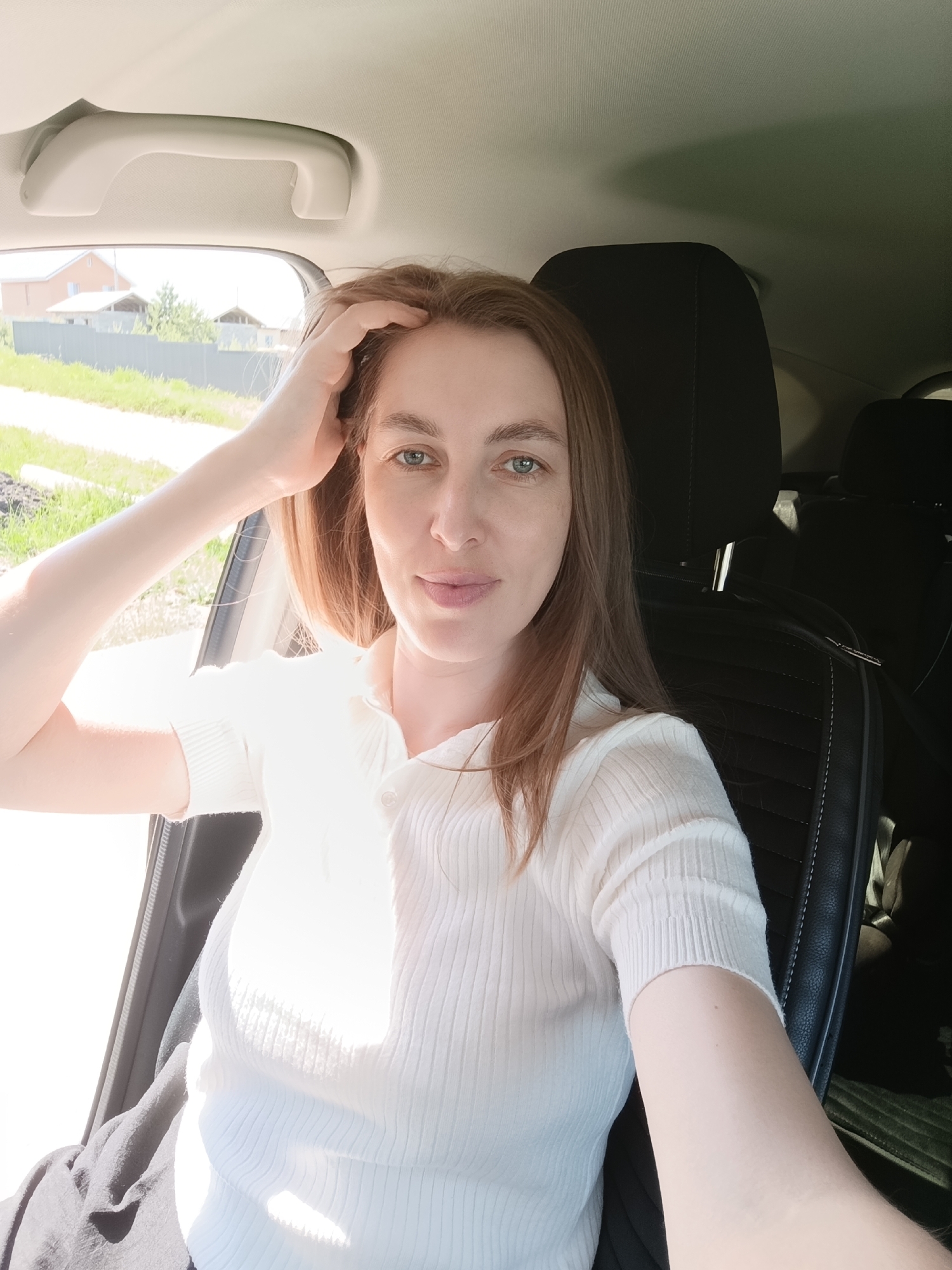 A woman with light brown hair and fair skin takes a selfie inside a car. She is wearing a white, short-sleeved ribbed shirt. Her left hand is resting on her head, and she is looking directly at the camera with a slight smile.
The car's interior is visible, including the black headrest, and the black seat with visible stitching. Outside the car window, a landscape can be seen with green grass and a grey fence. A house is visible in the distance.
Based on the available information, a specific location within the city of Chaikovsky, Russia cannot be determined.