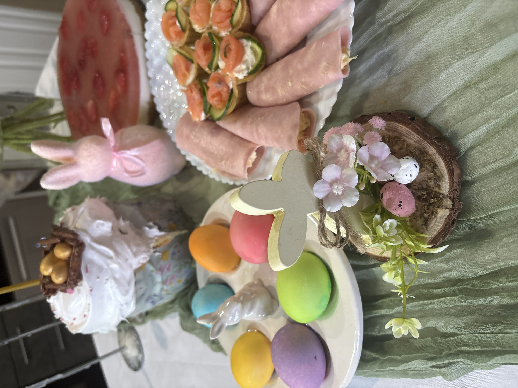 The image depicts an Easter-themed table setting. A white plate holds rolled ham and salmon appetizers with cucumber. Beside it, a pink bunny figurine and a watermelon-shaped decoration are visible. A cake with white frosting, decorated with a chocolate nest filled with golden eggs, sits alongside an egg holder containing pastel-colored Easter eggs and a small ceramic bunny. A wooden cross decorated with flowers rests on a wooden slice. A green linen tablecloth drapes beneath the items. The location is likely in Chaikovsky, Russia.