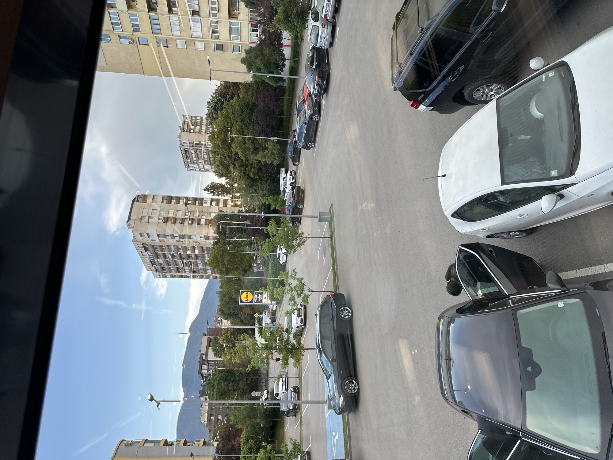 The image depicts a parking lot in Sofia, Bulgaria, with multiple cars parked and two women interacting near a dark sedan. The parking lot is surrounded by residential buildings and trees, with a mountain range visible in the background. The sky is partly cloudy, and it appears to be late afternoon or early evening.
In the foreground, a dark-colored sedan is parked in a marked parking spot, with its driver's side door and the rear passenger door open. A woman, wearing a dark jacket and jeans, is leaning into the open rear door, appearing to retrieve or place something inside. Her dark hair is pulled back in a ponytail. Another woman, wearing a lighter colored top and jeans, is standing to the left of the sedan, partially visible, with her hand on the open driver's side door.
Several other vehicles are parked in the lot, including a white hatchback, a black SUV, and various sedans. A blue handicapped parking symbol is visible on the ground near the dark sedan. A sign for "Lidl" is visible in the background, indicating a nearby supermarket. The buildings are multi-story apartment blocks, typical of urban Bulgarian architecture. The overall scene is that of an ordinary day in a residential area of Sofia.