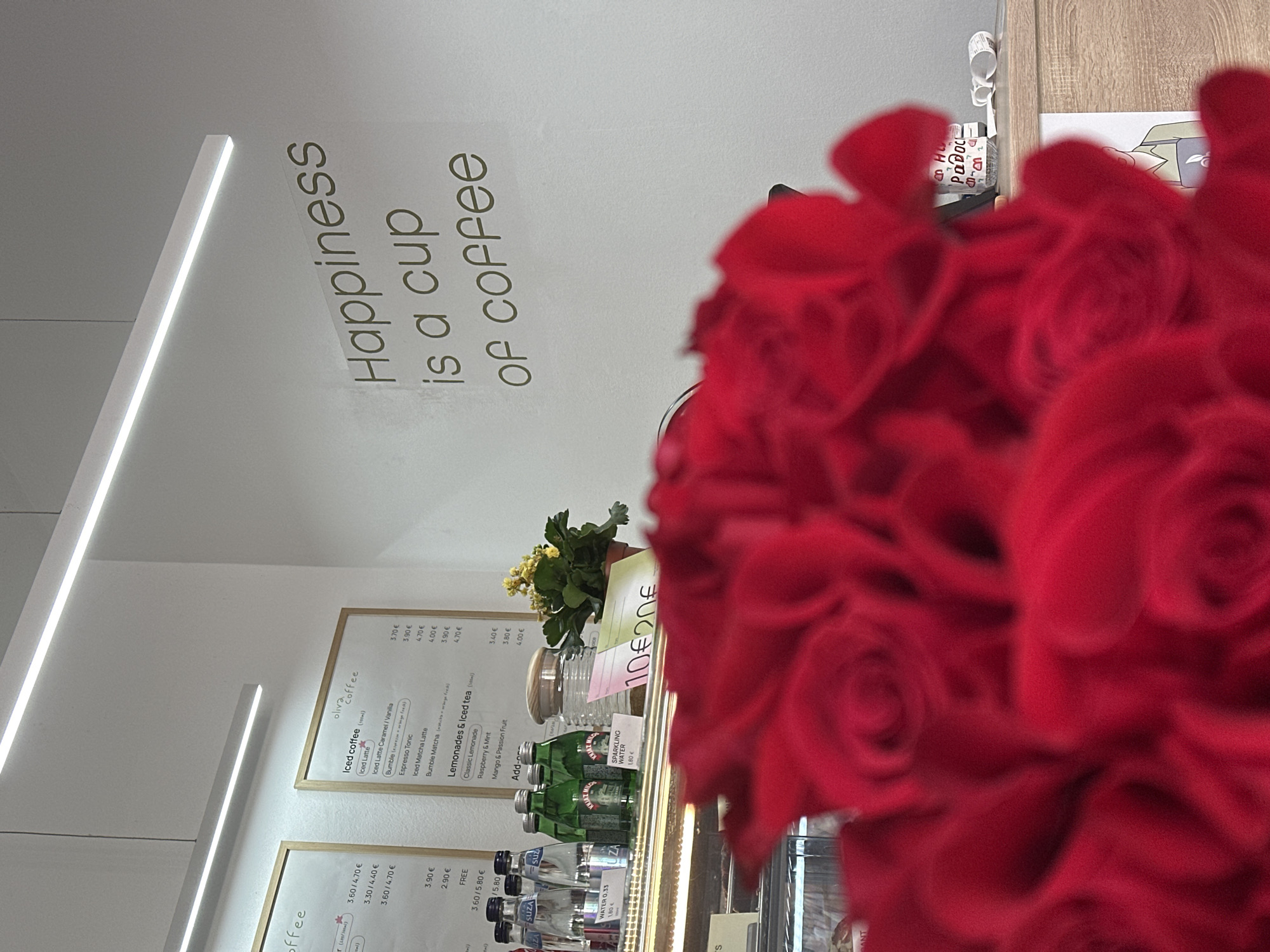 This indoor scene shows a coffee shop menu with a sign that reads "Happiness is a cup of coffee." The menu features iced coffee, lemonades, and iced tea. Prices are displayed in euros. In the foreground, a bouquet of red roses is partially obscuring the view. Below the menu are bottled beverages, including water and sparkling water. The location could be in Šušanj, Montenegro, based on context, though there are no explicit location identifiers in the image.