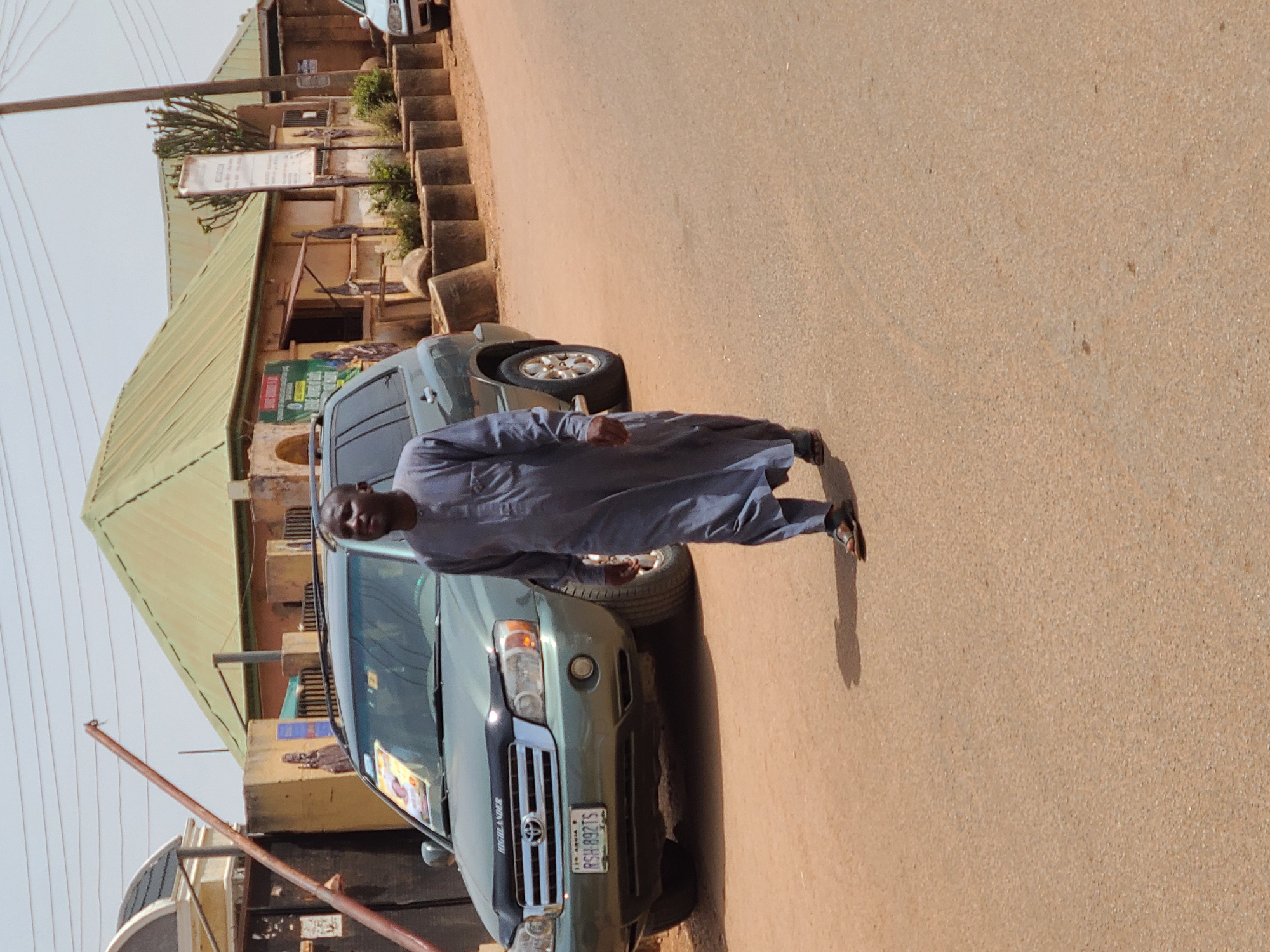 This image presents a daytime street scene in Jos, Plateau State, Nigeria, under a clear sky.
A dark-skinned adult male, dressed in a grey-blue long-sleeved tunic and trousers with dark sandals, walks in the central foreground towards the left. His shadow is cast prominently to his right.
To the left, a dark green, first-generation Toyota Highlander SUV is parked, partially facing the viewer. Its front bumper displays a Nigerian license plate, "Jos, Plateau State RSH-892TS," and a poster is affixed to its windshield. Behind the vehicle, a light-colored building features a dark, ornate roof over a gate, adjacent to another building with a light olive-green corrugated metal roof and light brown walls. A reddish-brown metal pole extends diagonally upwards from the far left.
On the right side of the image, a row of single-story buildings with light olive-green corrugated metal roofs and light brown walls lines the road. In front of these buildings, several concrete cylindrical planters or bollards are arranged, some containing small plants. Various signs are affixed to the buildings, including a prominent white rectangular sign on a pole. A large, multi-branched succulent plant is visible further back. The front portion of a light-colored car is partially visible on the far right. The ground throughout the scene is a light brown, unpaved or dirt road surface.