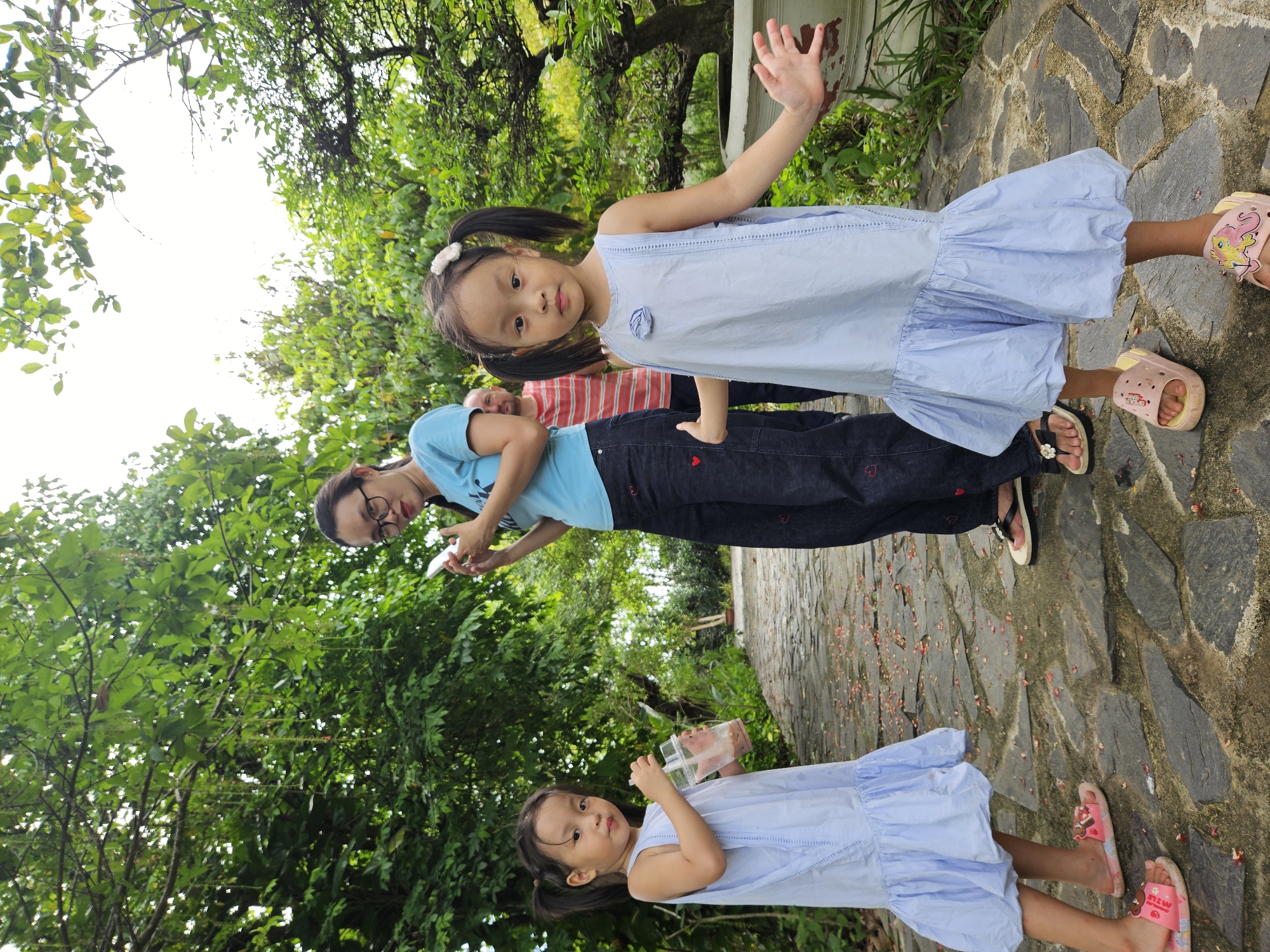 A family is posing for a photo in an outdoor garden setting, possibly in Da Nang, Vietnam. In the foreground, two young girls stand on a stone pathway. The girl on the left, wearing a light blue dress and pink sandals, holds a plastic cup with a straw. The girl on the right, also in a light blue dress and pink sandals, has her arms outstretched. Behind them, a woman in a blue t-shirt and dark patterned pants holds a smartphone. A man in a striped shirt is visible behind the woman. The pathway is paved with irregular gray stones and is littered with small red petals. Lush green trees and foliage surround the area, with dappled sunlight filtering through the leaves.