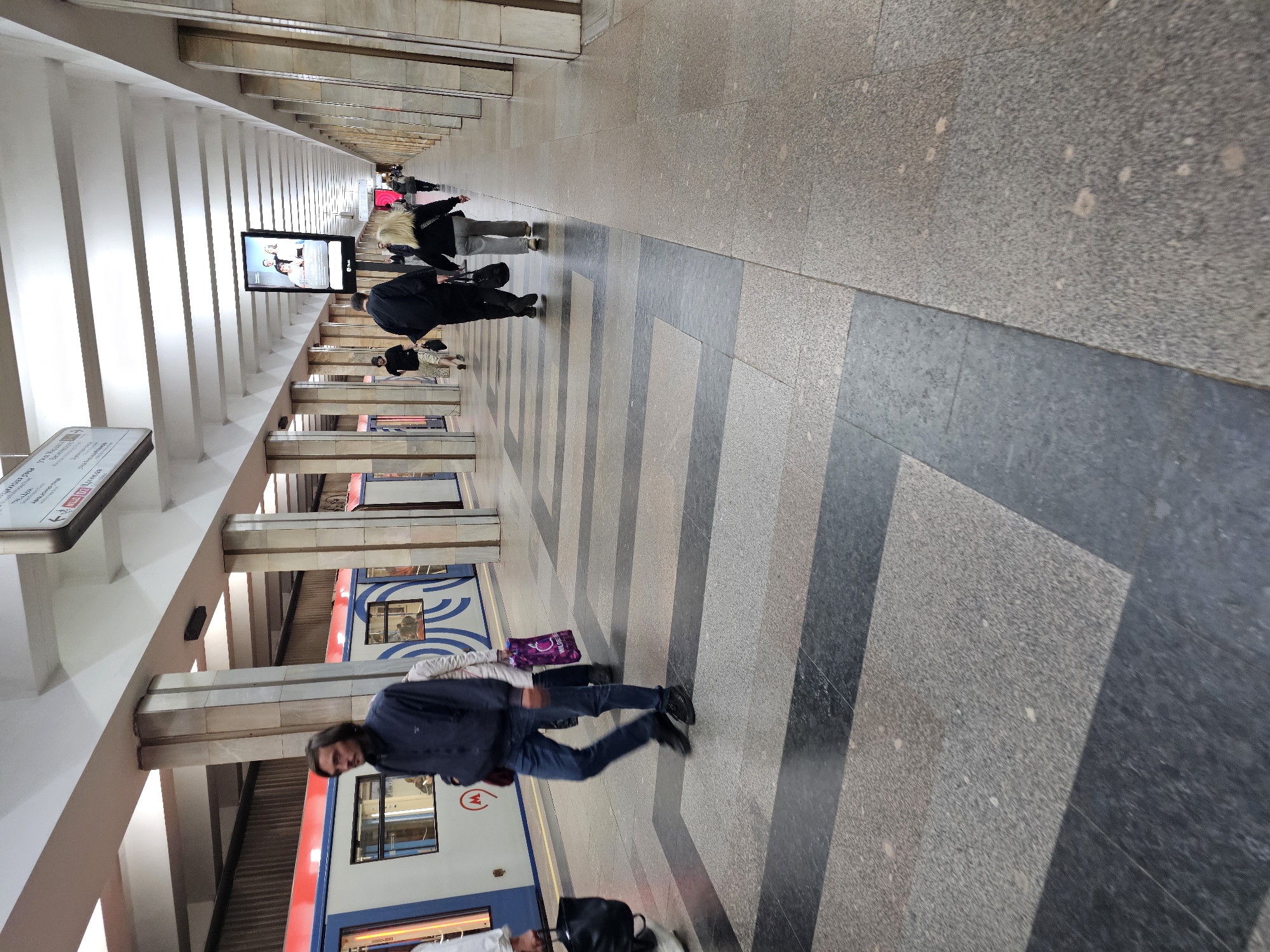 An outdoor metro station platform features a train with red and white carriages on the left side of the frame. In the center, a man with long dark hair and a blue jacket walks away from the camera. He wears blue jeans and black shoes. To his right, a person in a white jacket and dark pants carries a purple shopping bag. Further down the platform, several other individuals are visible walking away from the camera. The platform is paved with large grey and dark grey rectangular tiles. A row of large, fluted concrete pillars runs down the right side of the platform, supporting an overhead ceiling structure. Above the pillars, screens display advertisements and information. The scene appears to be in a metro station.