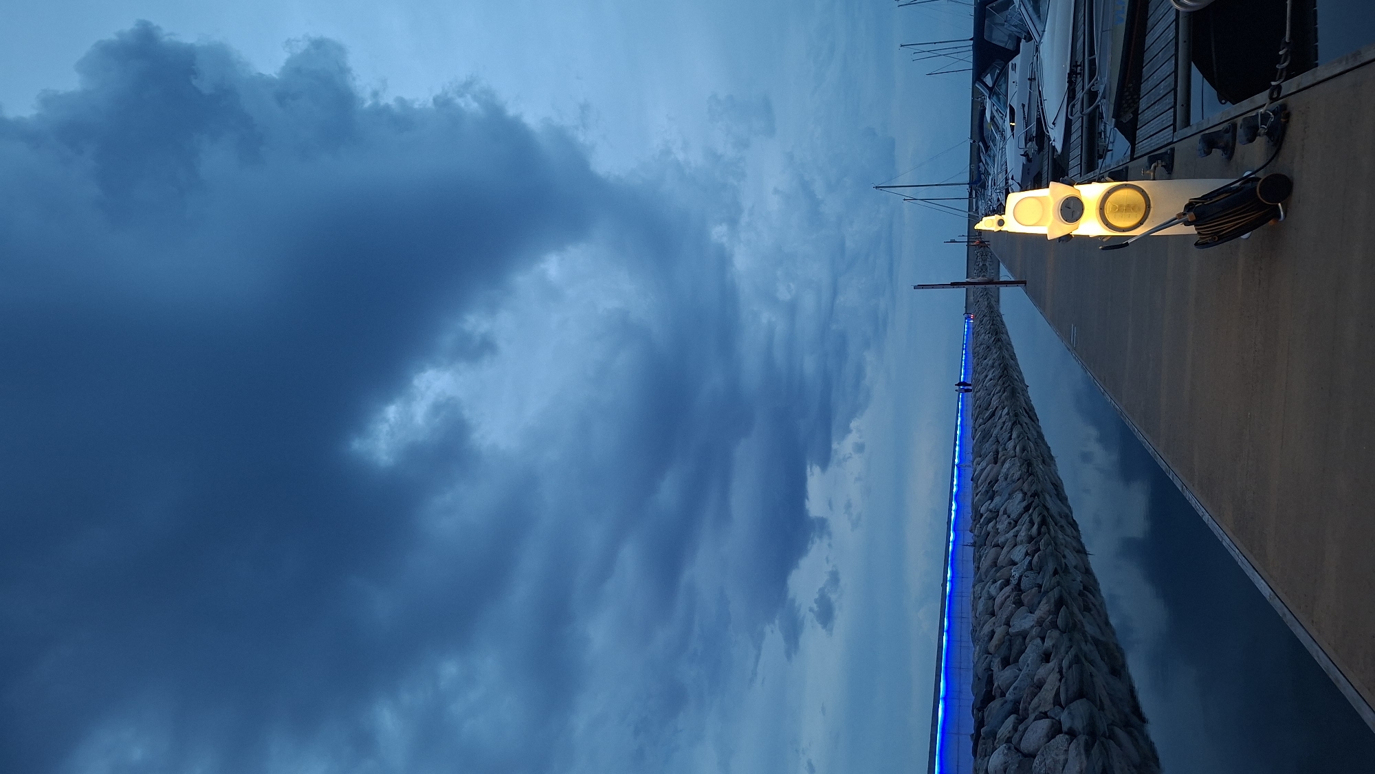 This is a vertical, eye-level, medium shot of a harbor walkway at dusk, located in Tallinn, Estonia. The scene is dominated by dramatic, dark blue clouds filling the upper left portion of the frame, suggesting an impending storm or the transition from day to night. The lower right of the image shows a concrete walkway alongside a calm body of water. Along the edge of the walkway, a stone barrier is illuminated with bright blue LED lights, creating a striking contrast with the dark sky and water. Further down the walkway, a series of three modern, circular yellow lights are mounted on poles, casting a warm glow. A black and silver piece of equipment, possibly a speaker or a light fixture, is attached to the rightmost yellow light. Beyond the walkway, several sailboats are docked, their masts and rigging visible against the dimming sky. The overall mood is atmospheric and slightly dramatic, amplified by the contrasting lights and the brooding clouds. No visible text is present in the image. The time of day is clearly dusk, given the low light conditions and the illumination from the artificial lights.