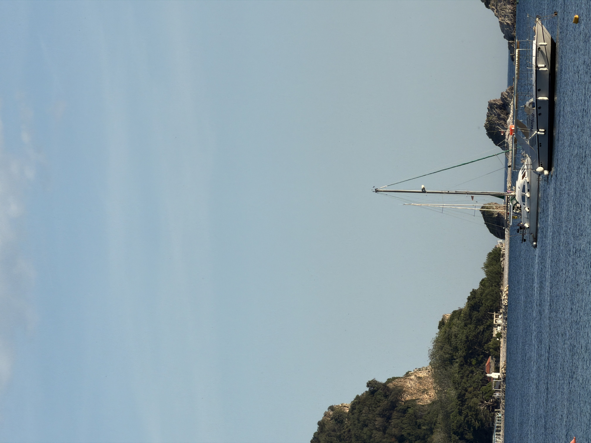 The image shows a coastal scene in Budva, Montenegro, featuring two sailboats anchored in the water near a rocky, tree-covered coastline. The sky is clear and blue. The boats are different sizes, with the larger one appearing to be a motor yacht. In the distance, other rocky formations are visible. There are houses and buildings along the coastline. The overall impression is that of a calm, sunny day at a popular coastal location.