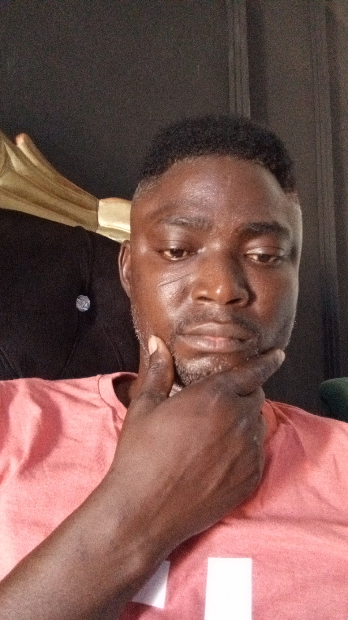 A man with dark skin, short dark hair, and a salt-and-pepper beard is the subject of a close-up photograph. He is wearing a light pink shirt. His right hand is raised to his chin in a contemplative pose. The background consists of a black chair with a tufted back and a gold-colored headrest. The location in the city of Sabon Tasha, Nigeria is not evident from the image.