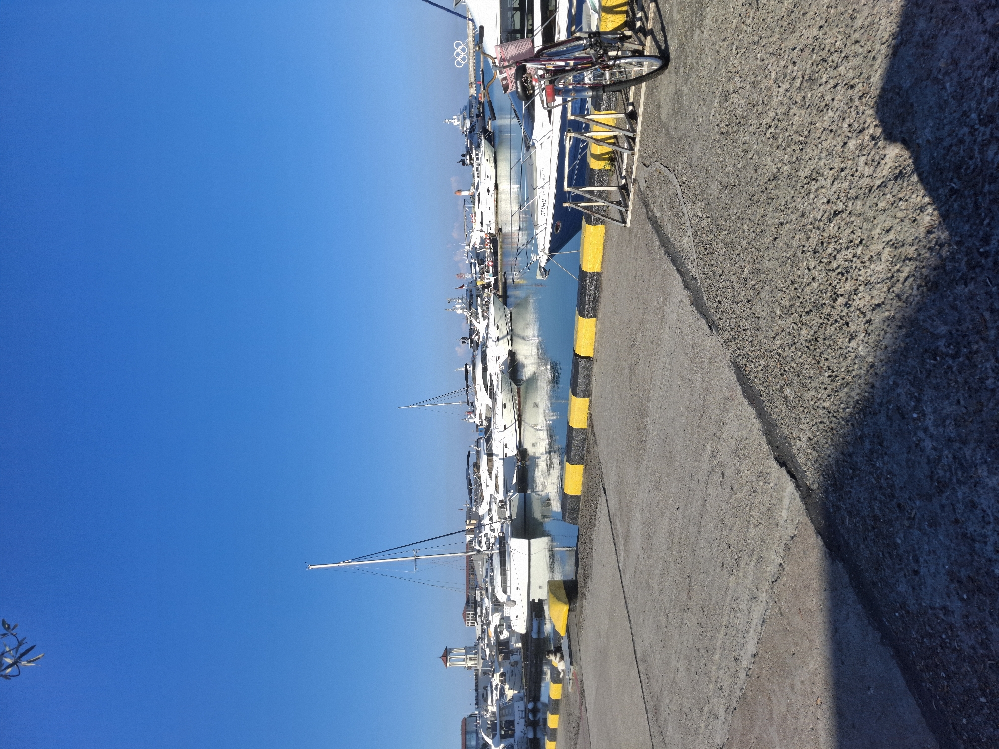 The image depicts a marina in Sochi, Russia, likely located near the Olympic Park, evidenced by the presence of the Olympic rings sculpture in the background. The foreground features a concrete pier with distinct black and yellow painted boundaries. A bicycle is parked on the pier, leaning against a bicycle rack. In the mid-ground, a fleet of white yachts and sailboats are moored in the clear blue water. The sky is a vibrant, cloudless blue. The overall scene conveys a sense of leisure and a bright, sunny day at the waterfront.